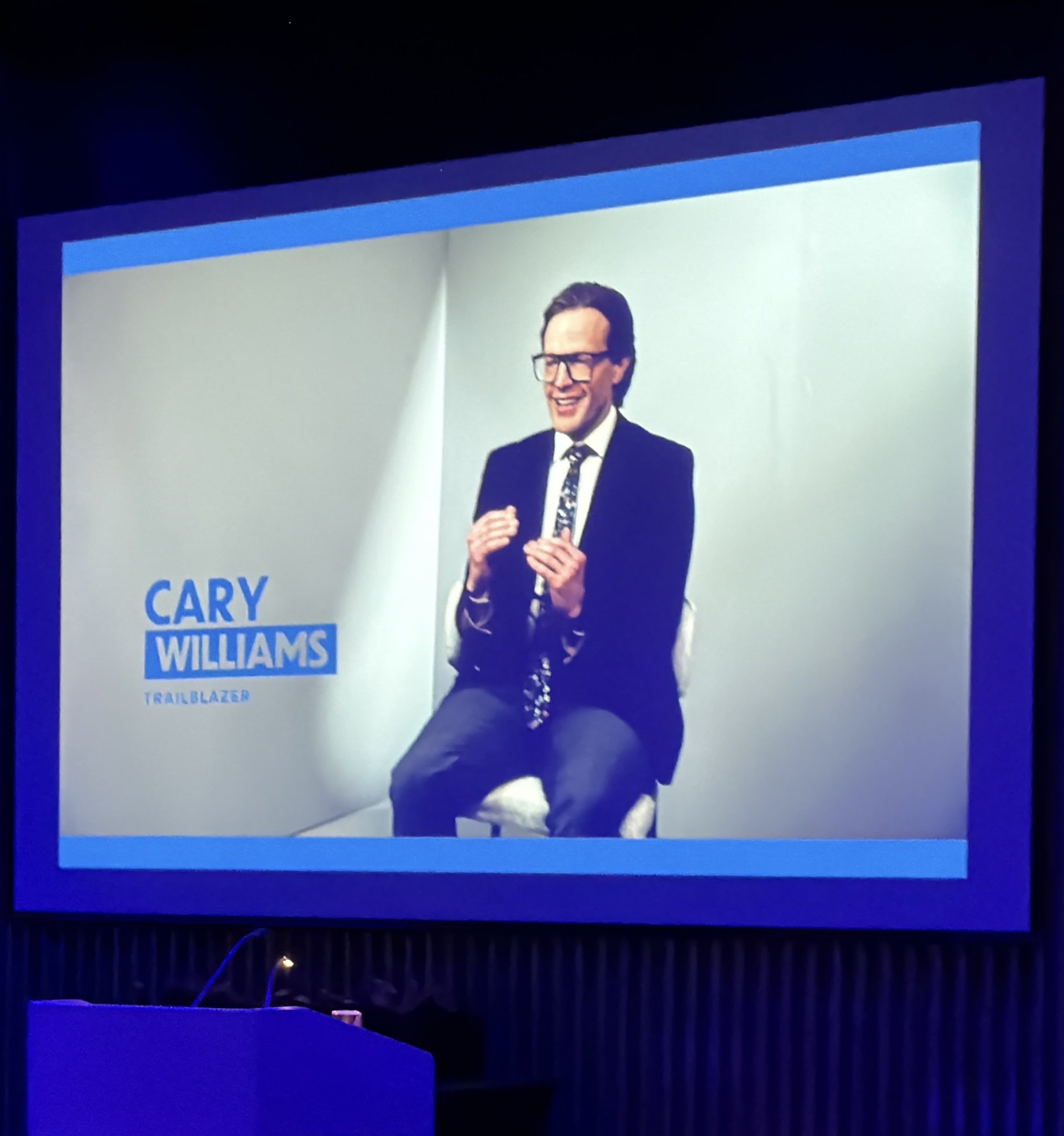 A man in a suit and glasses speaking at an event with a large screen behind him displaying his name, 'Cary WILLIAMS,' and other text, under purple and blue lighting.