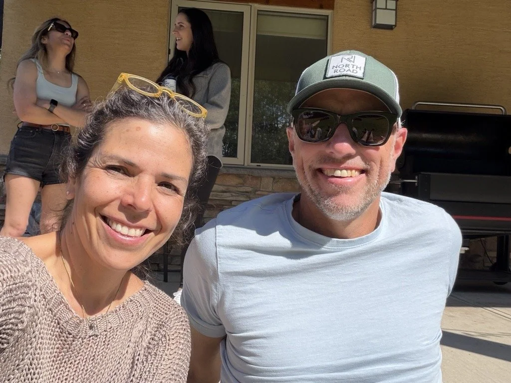Two women and a man smiling for a selfie outside a house, with two other women talking in the background near a window and a grill.