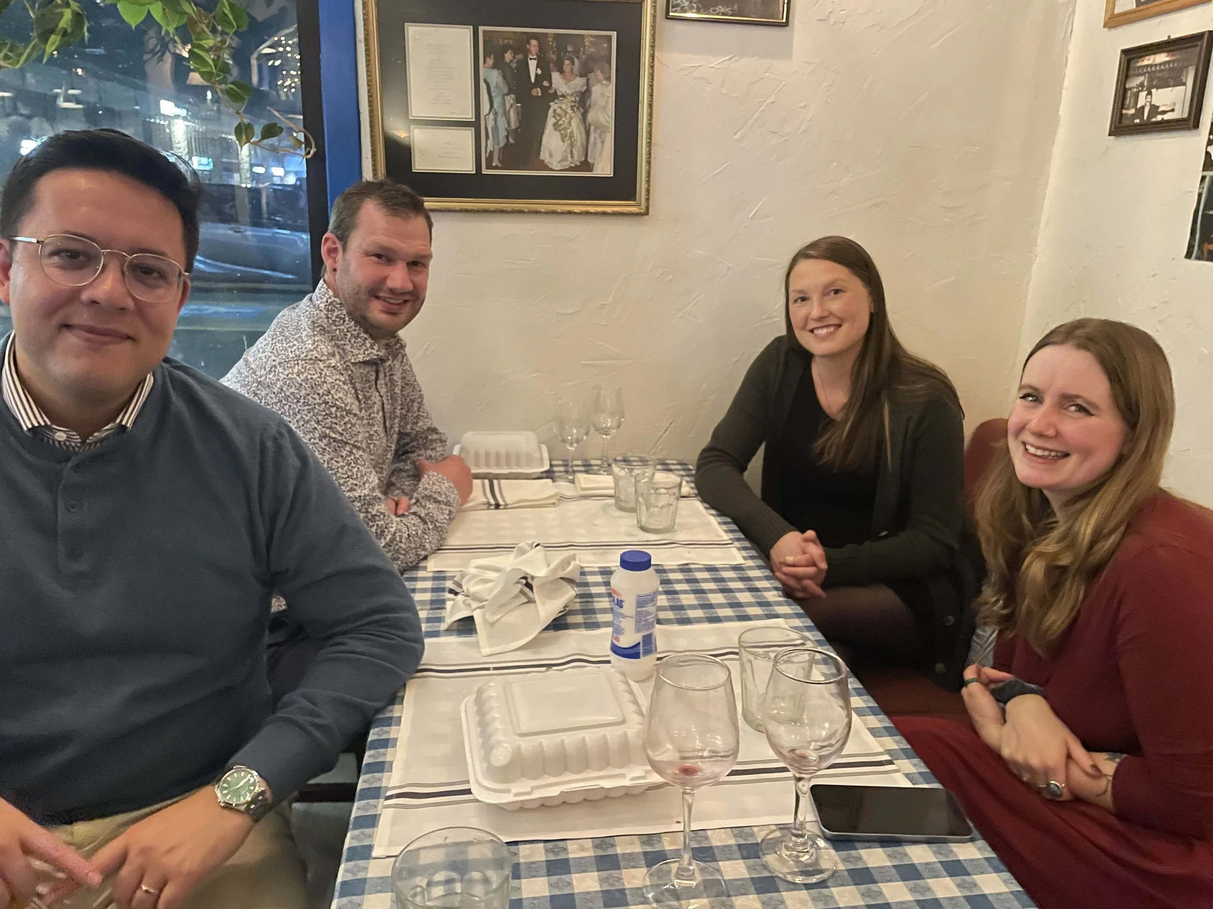 Four people sitting at a table in a restaurant, smiling at the camera. The table has a blue and white checkered tablecloth, empty glasses, a white takeout container, napkins, and a drink bottle. The background features framed pictures on a beige wall