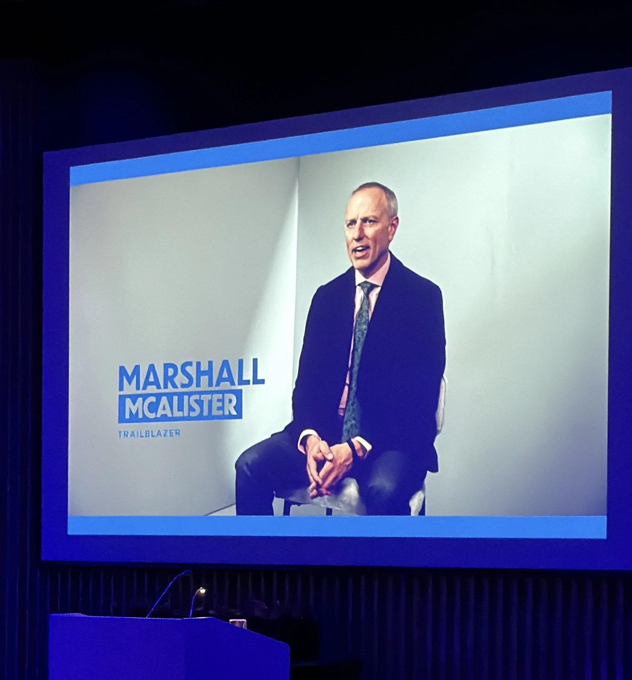 A person in a suit is speaking at a conference, with a presentation slide that reads 'MARSHALL MCCLISTER.' The slide has a blue and white color scheme, and the person appears to be mid-sentence, seated on a stage with a black background.