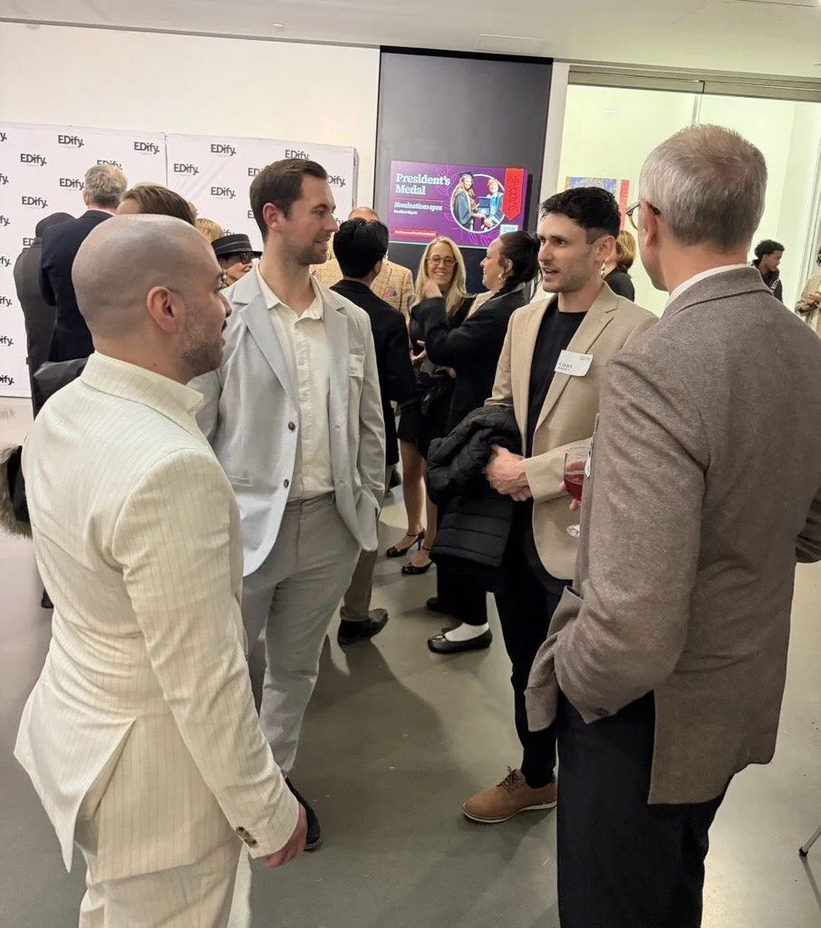 Group of people in business attire having a conversation at an event with a banner and digital screen in the background.
