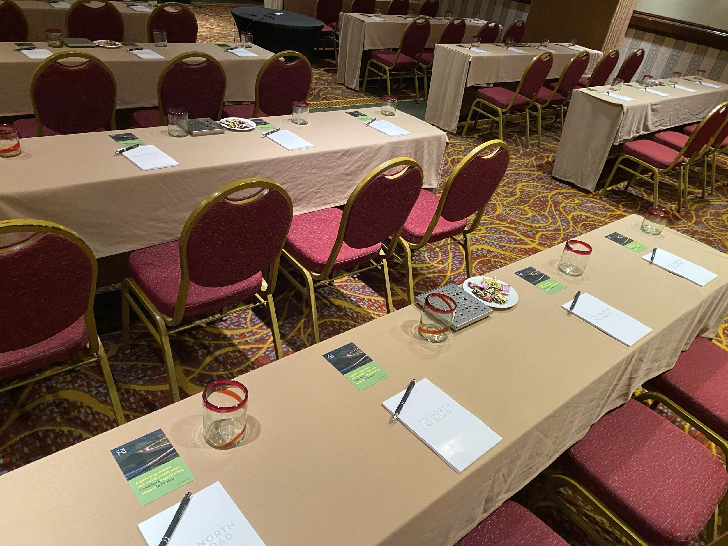 Conference room with long tables covered in beige tablecloths and red chairs. Each table has notebooks, pens, glasses with red rims, and green pamphlets. The room has patterned carpet and warm lighting.
