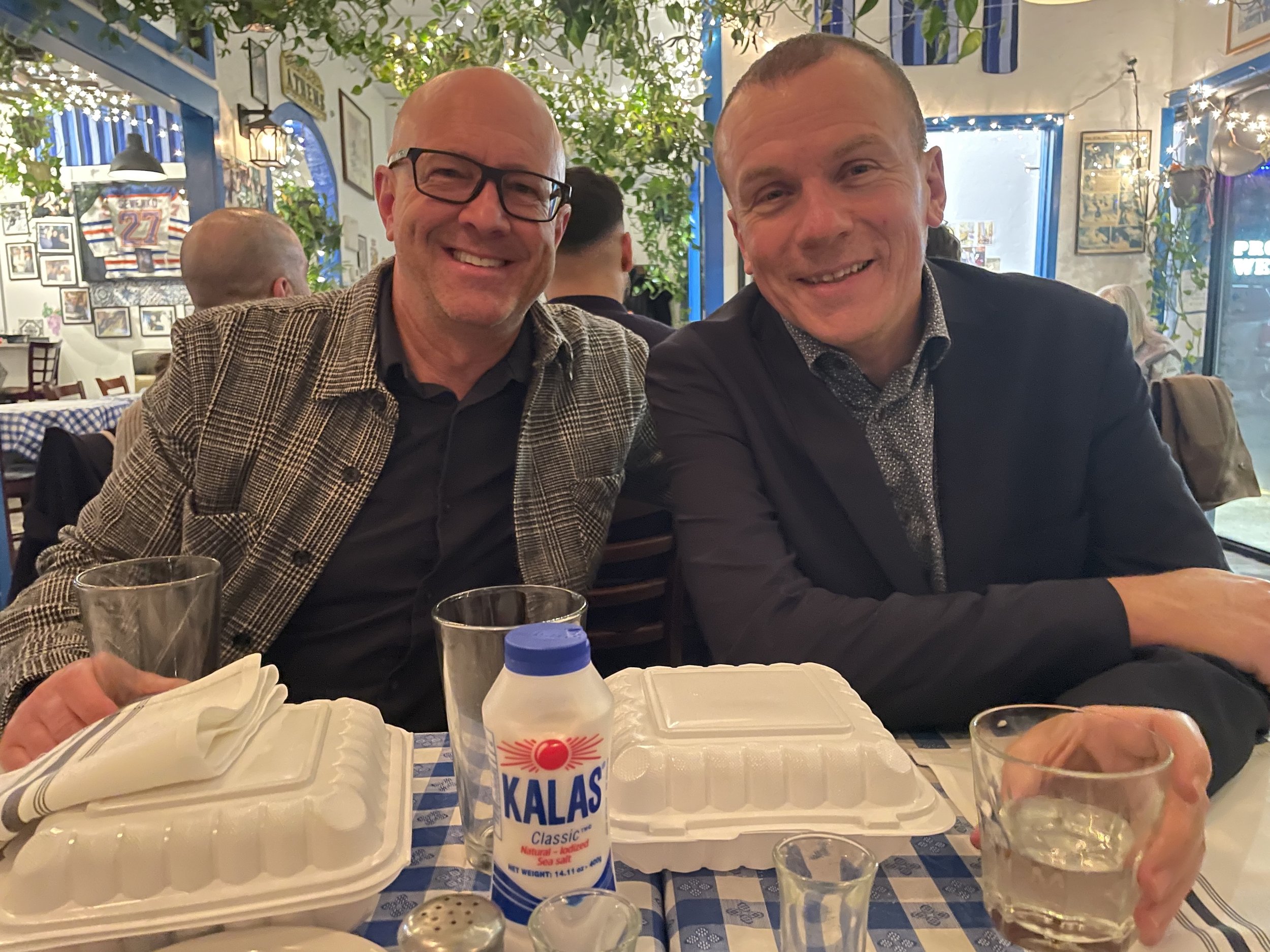 Two men sitting at a table inside a restaurant, smiling at the camera. The table has takeout containers, glasses, and a bottle of Kalas salt. The restaurant decor includes framed photos, string lights, and greenery.