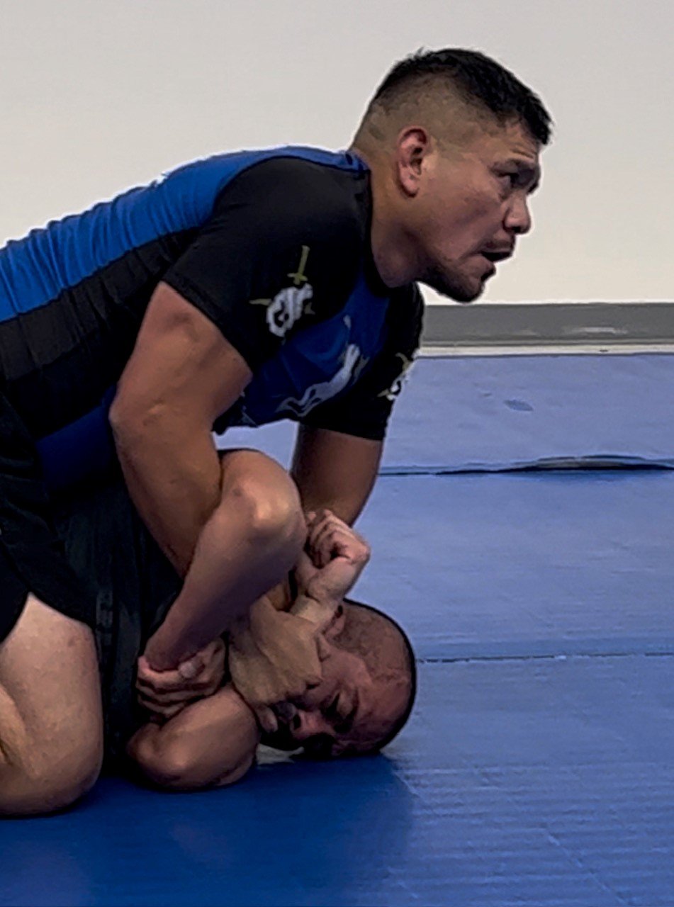 Two men practicing Brazilian Jiu-Jitsu on a blue mat, Professor Luke Harwood in a dominant position, holding the other in a chokehold.
