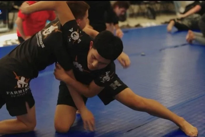 Two boys practicing Brazilian Jiu-Jitsu, one boy in black rash guard and shorts on top, the other in black rash guard and shorts on the bottom, with others practicing in the background.