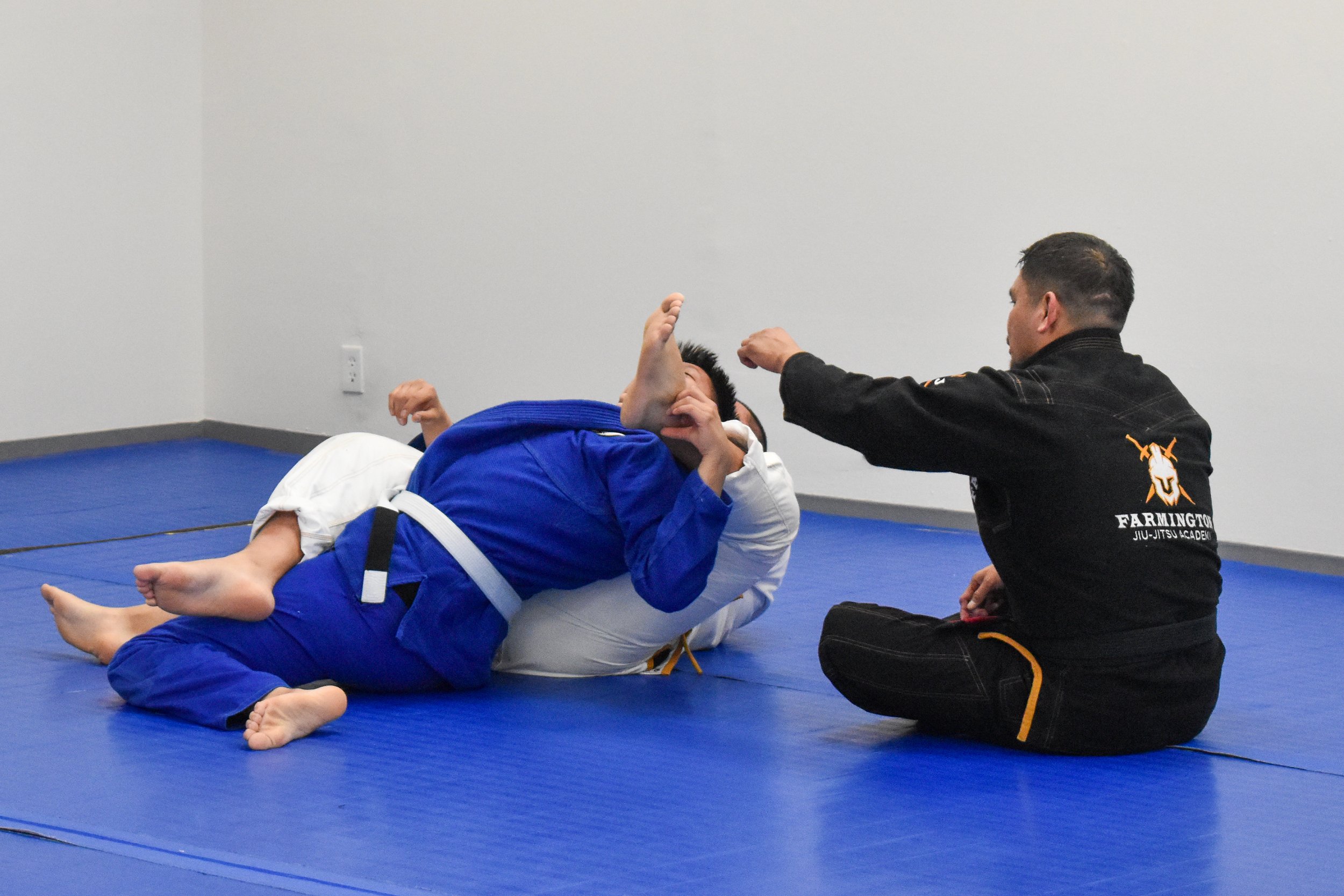Two Brazilian Jiu-Jitsu practitioners, one in a white gi and one in a blue gi, on a blue mat during training, with professor Luke Harwood observing and signaling from nearby.