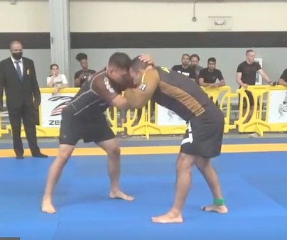 Two male grapplers competing, wrestling with each other, with spectators and officials watching in the background.
