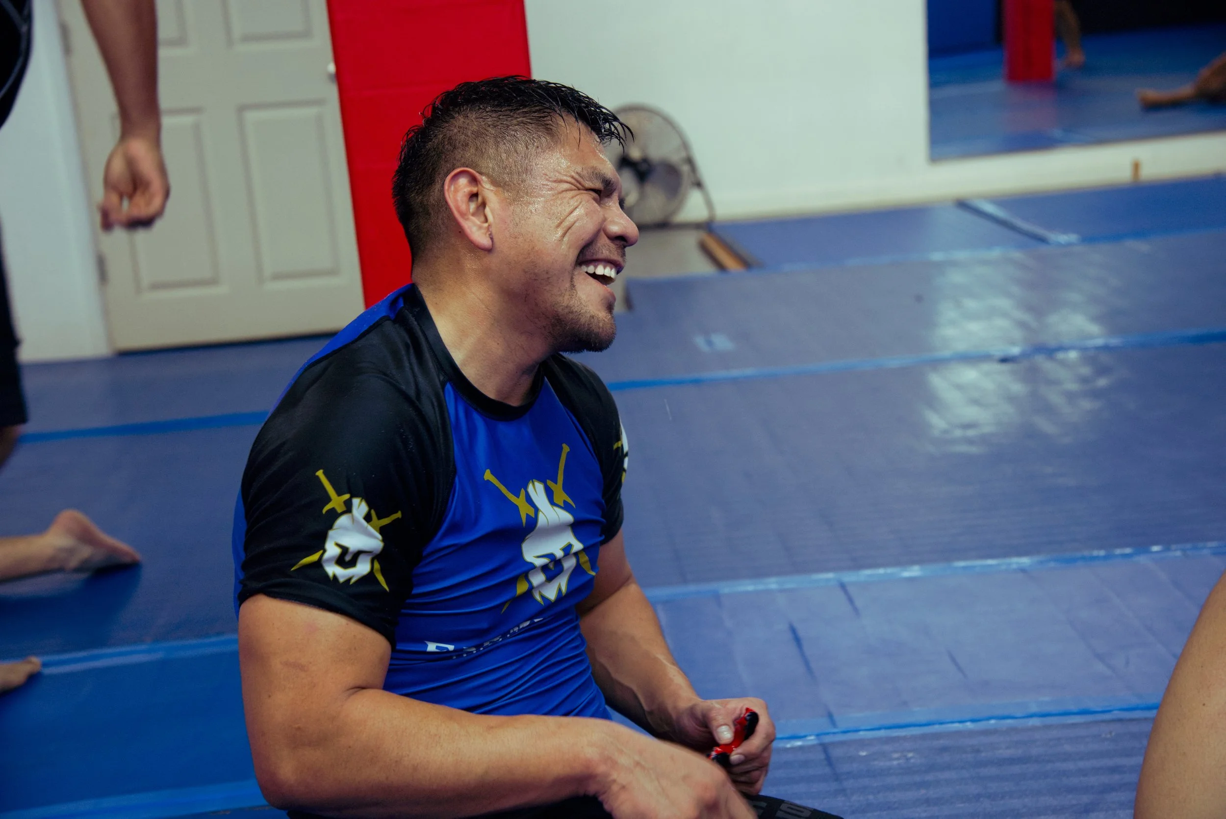 Man smiling and laughing while sitting on a padded blue floor in a gym, wearing a blue and black athletic shirt, with a red and white wall and a door in the background.