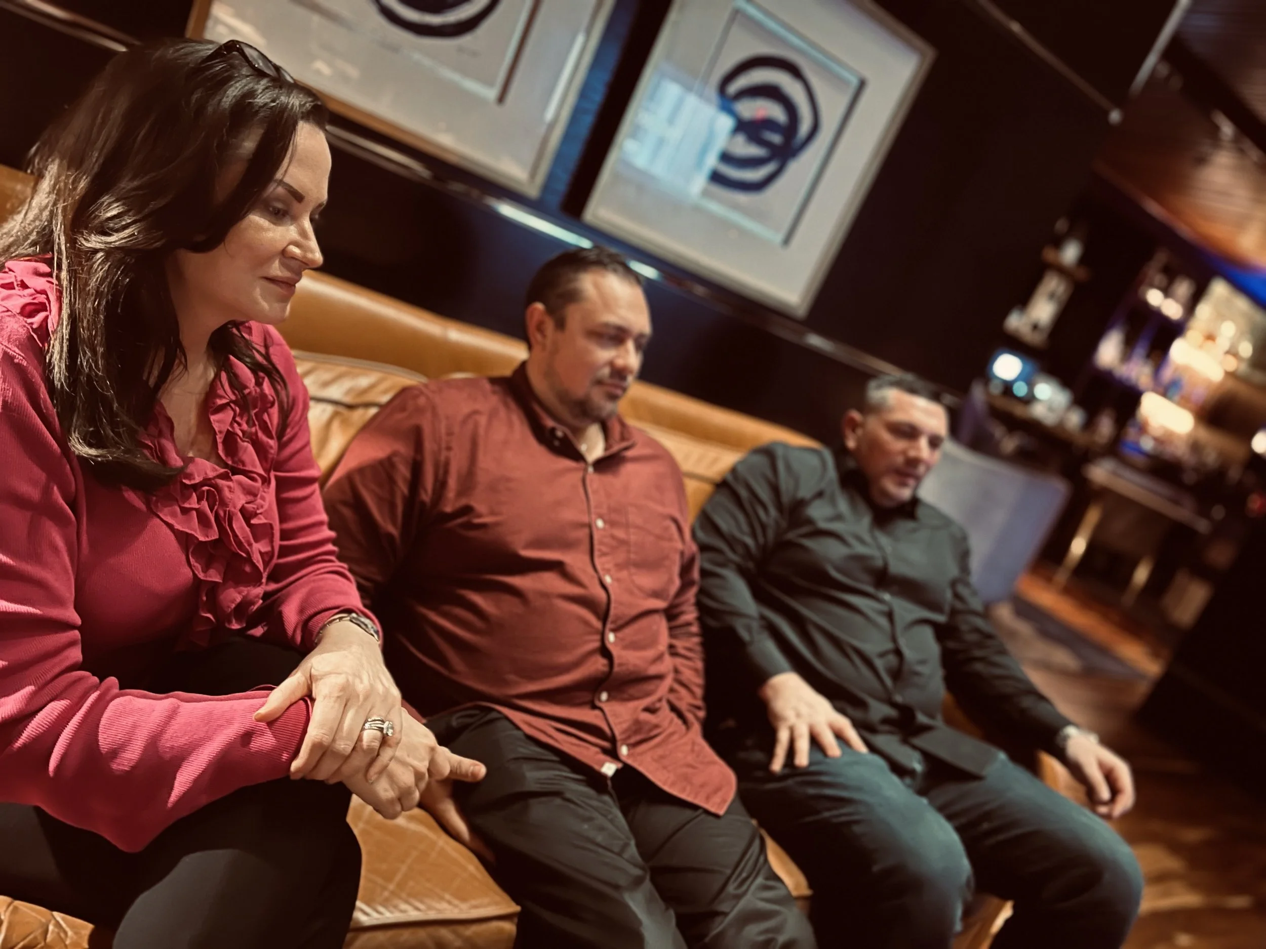 Three people sitting on a brown leather bench in a dimly lit bar or restaurant, engaged in conversation. The woman on the left has dark hair, wears a red ruffled blouse, and has her hands clasped in her lap. The man in the middle wears a reddish-brow