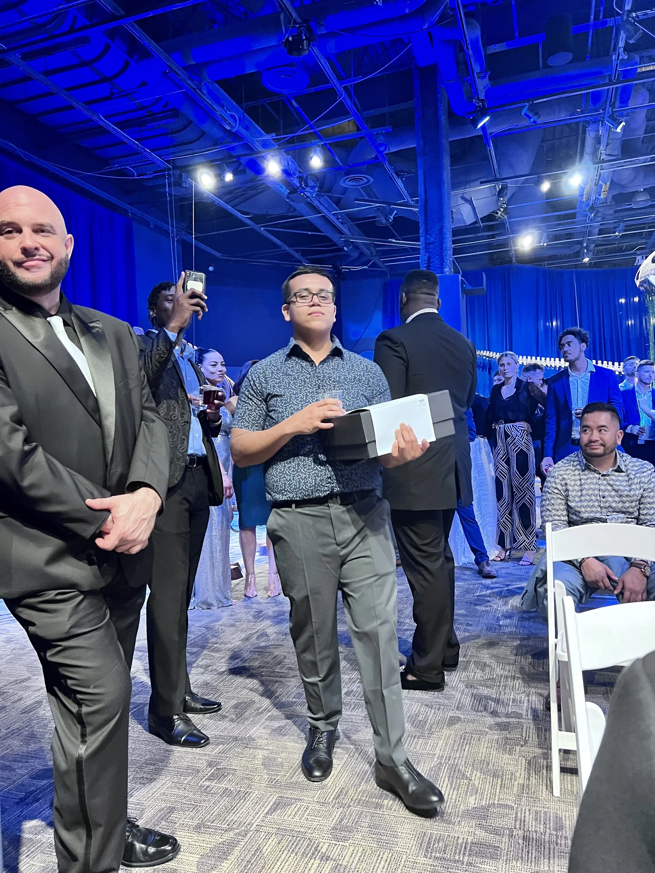 At an indoor event, a young man in a patterned shirt holds a box and a drink and walks confidently while others are gathered around, some taking photos or watching. The event has blue lighting and a high ceiling with visible metal beams.