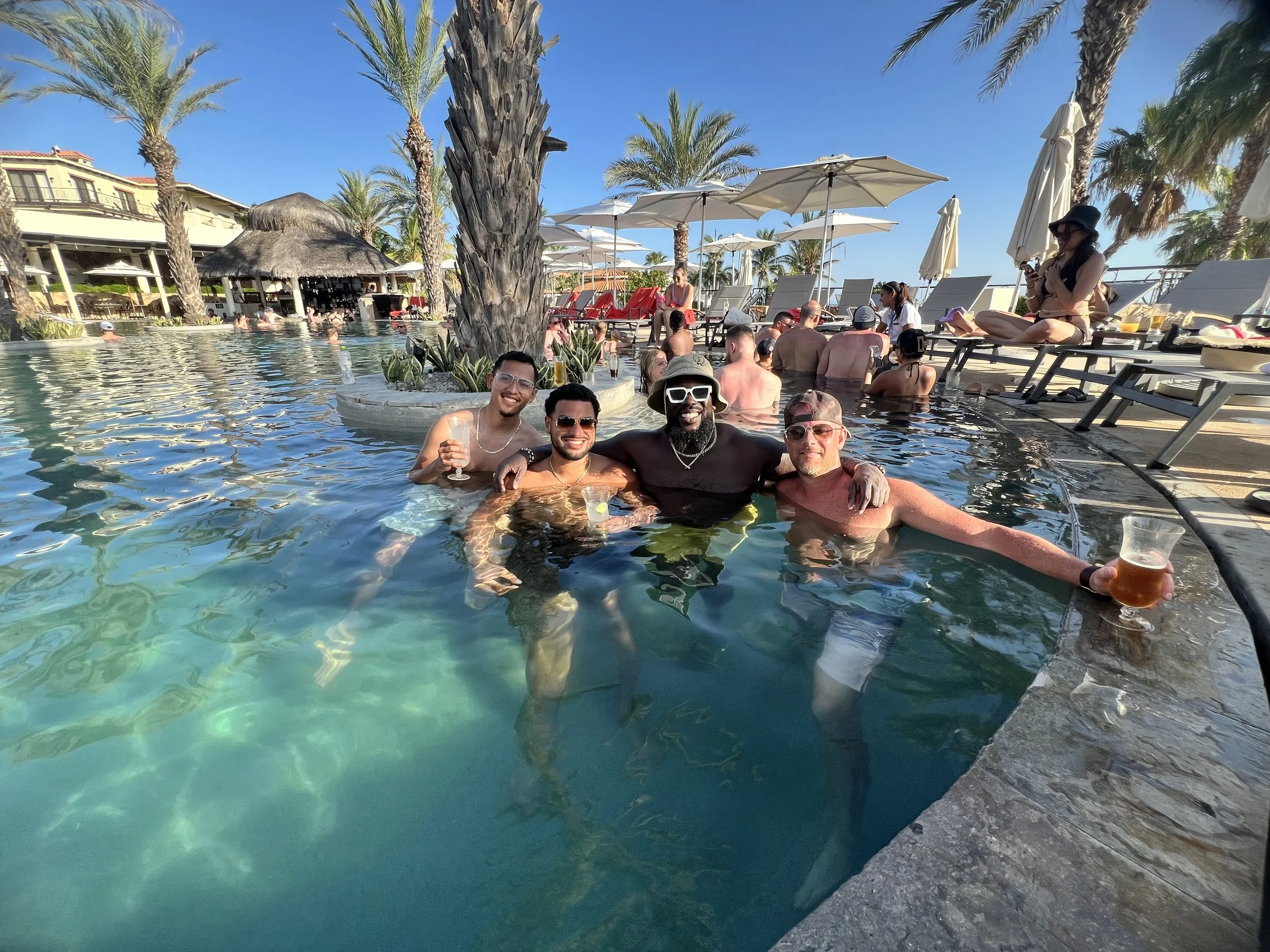 Four men smiling and enjoying drinks in a swimming pool at a lively resort, with palm trees and lounge chairs in the background, during daytime.