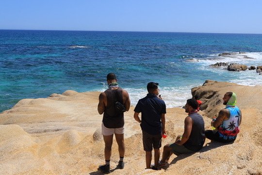 Four people sitting and standing on a rocky shoreline by the ocean, with waves and a clear blue sky in the background.