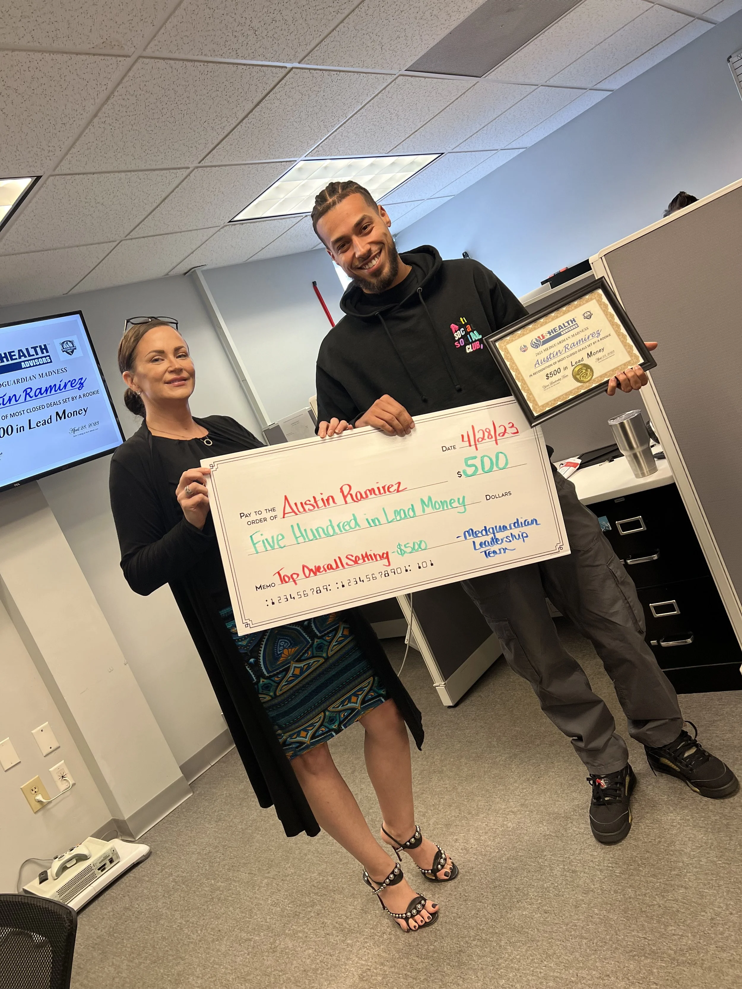 A man and woman are smiling and holding a large check and a framed certificate. The check is made out to Austin Ramirez for $500, dated April 28, 2023, and is from the Medguardian Leadership Team for a Top Overall Setting award. The woman is wearing 