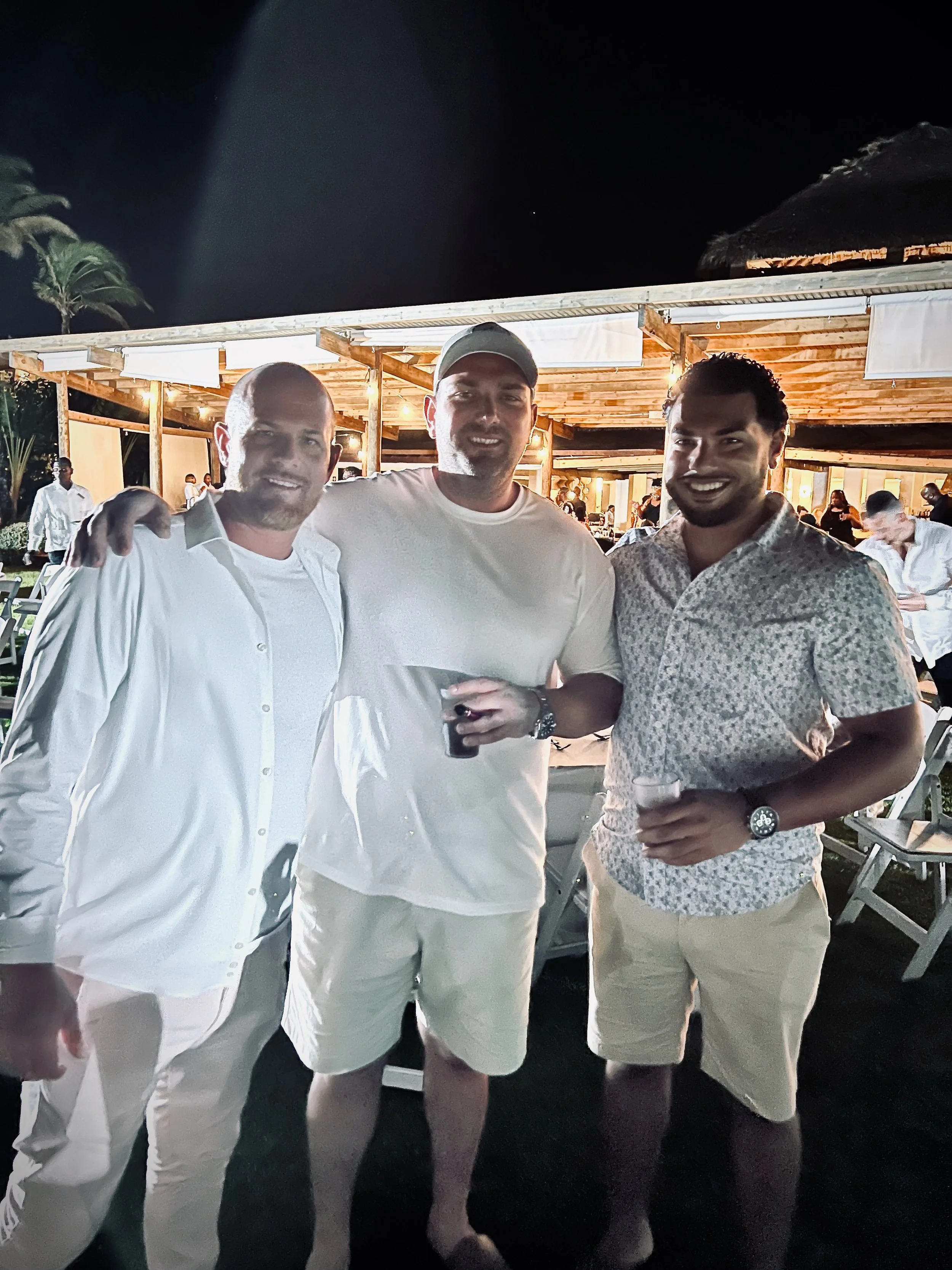 Three men standing together at night, smiling, at an outdoor event with a wooden pavilion and tables in the background, each holding a drink.