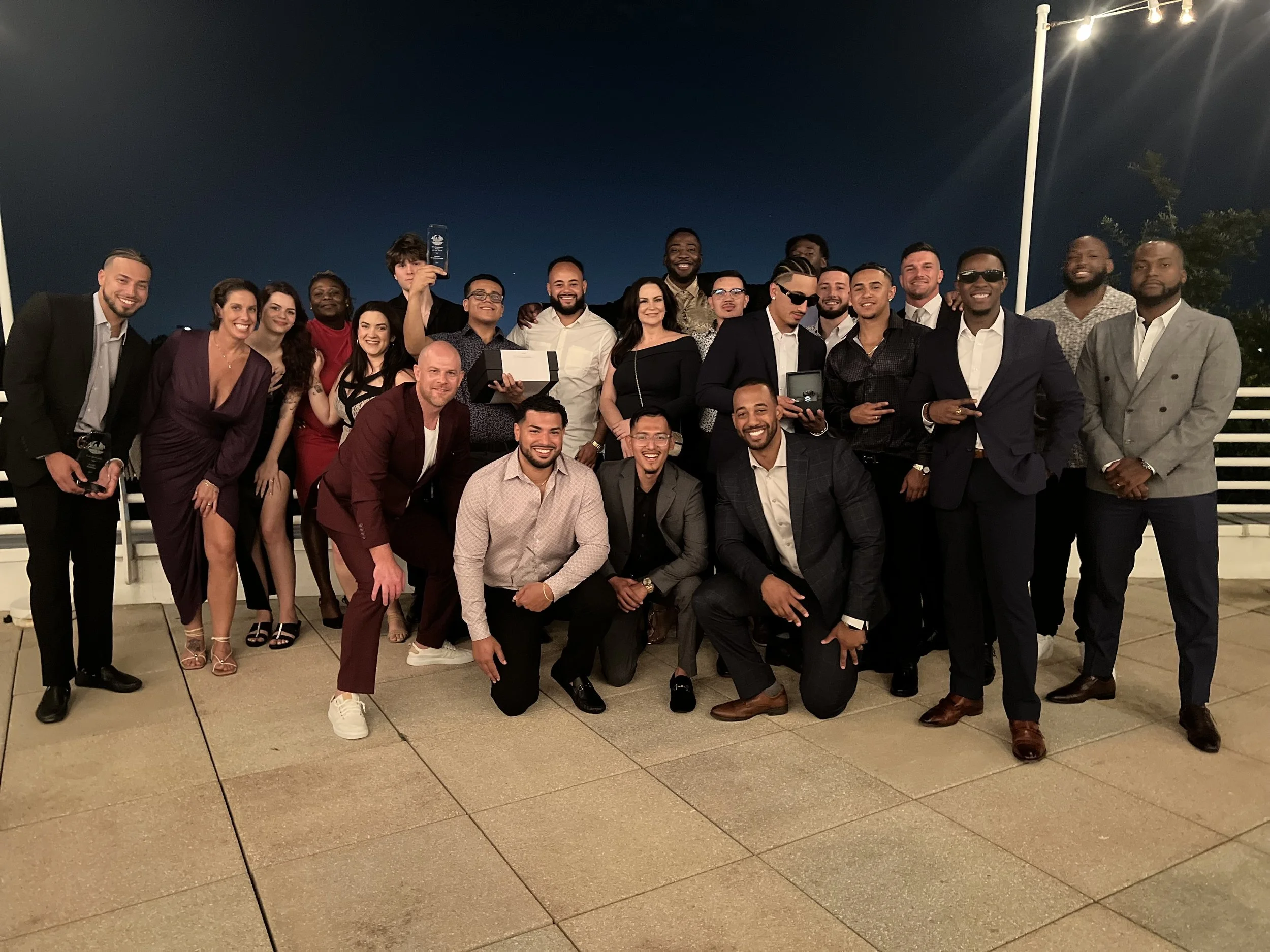 Group of about 24 young adults dressed in formal attire, posing together on a rooftop at night, celebrating an event with trees and string lights in the background.