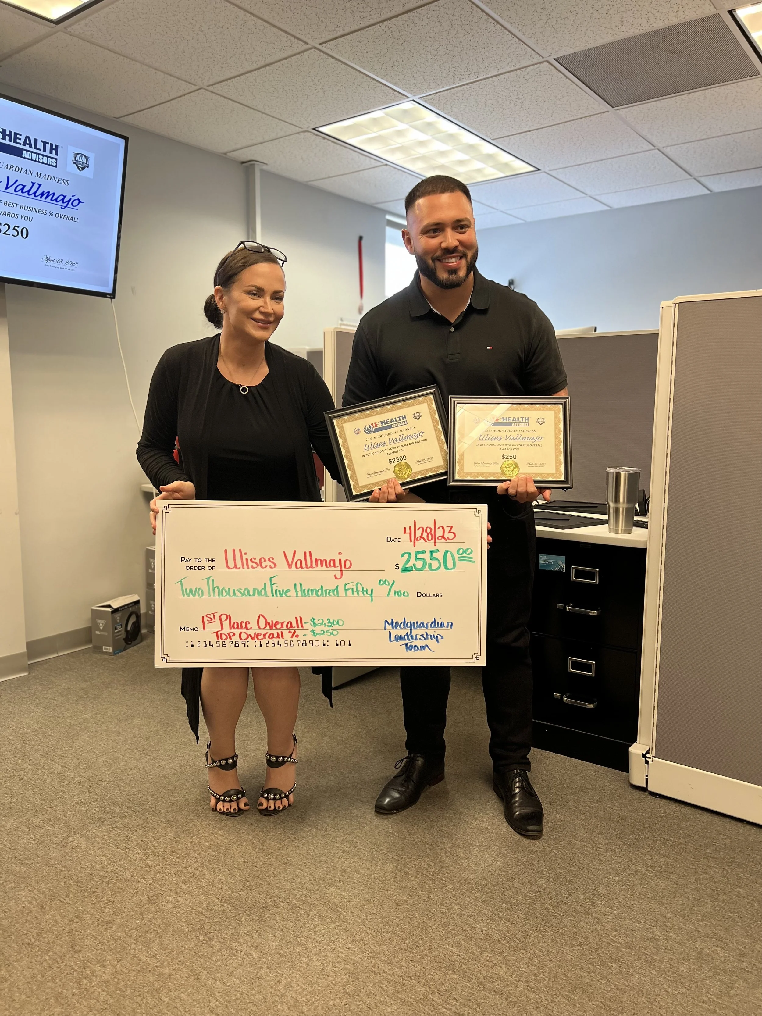 Two people, a woman and a man, standing in an office holding awards and a large check, celebrating an achievement. The woman is wearing black heels and a black dress, and the man is dressed in black pants and a black polo shirt. They are smiling and 