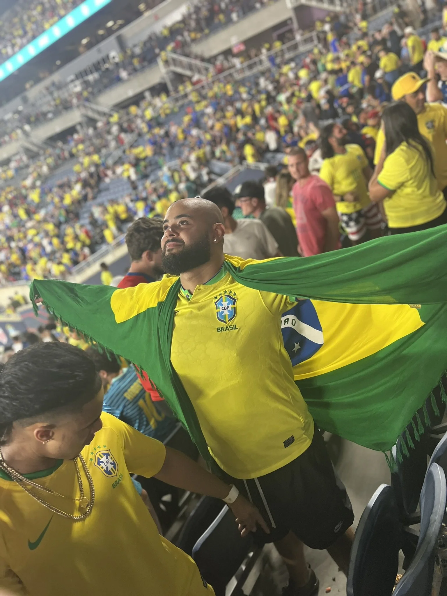 A man in a yellow Brazil soccer jersey holding a Brazilian flag and holding hands with a young boy in a yellow shirt at a crowded stadium.
