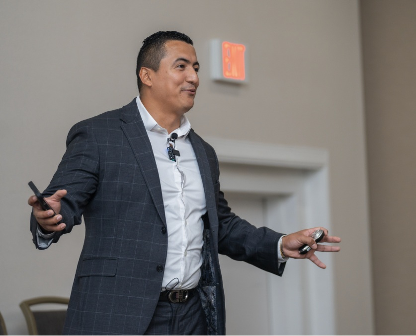 A man in a dark checkered suit presenting in a conference room with light-colored walls, holding a remote and a laser pointer, with an exit sign in the background.