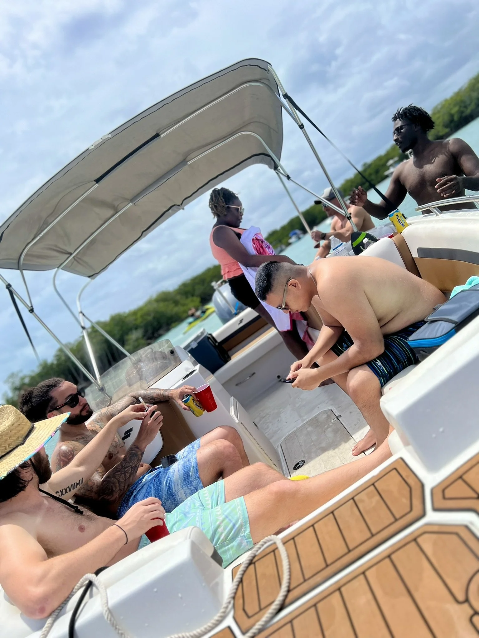 Group of people on a boat near water, some sitting and some standing, enjoying a sunny day. Some are shirtless, some wearing sunglasses, surrounded by trees and a cloudy sky.