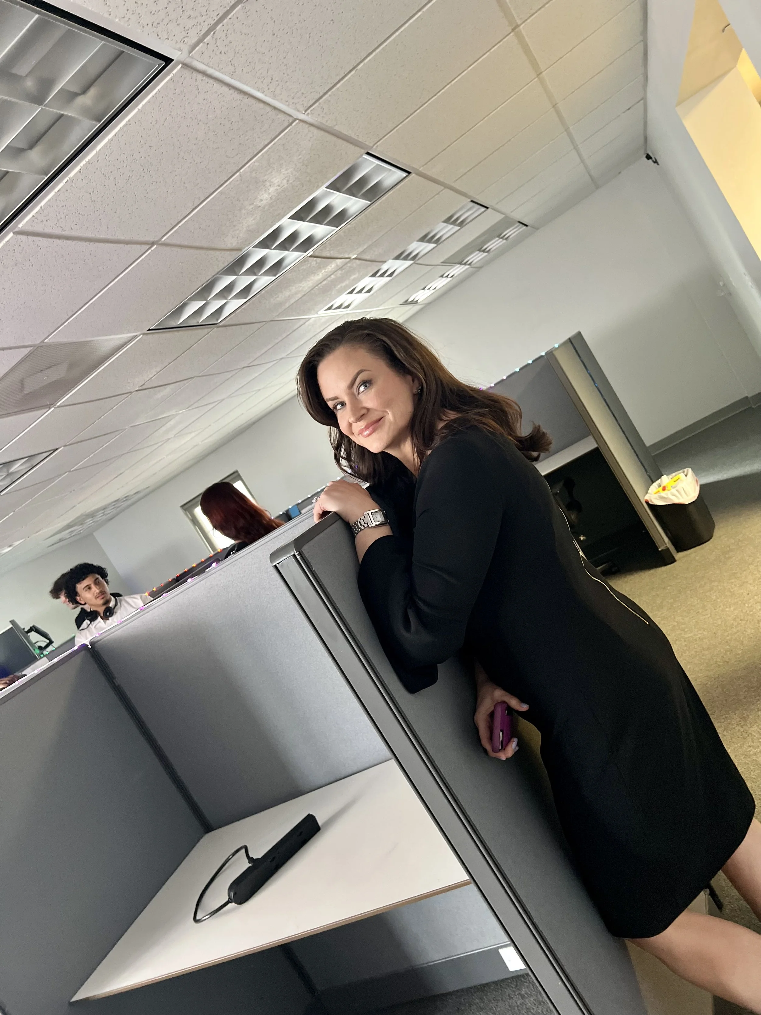 A woman in a black dress leaning on a cubicle wall in an office, smiling at the camera, with office coworkers working in the background.