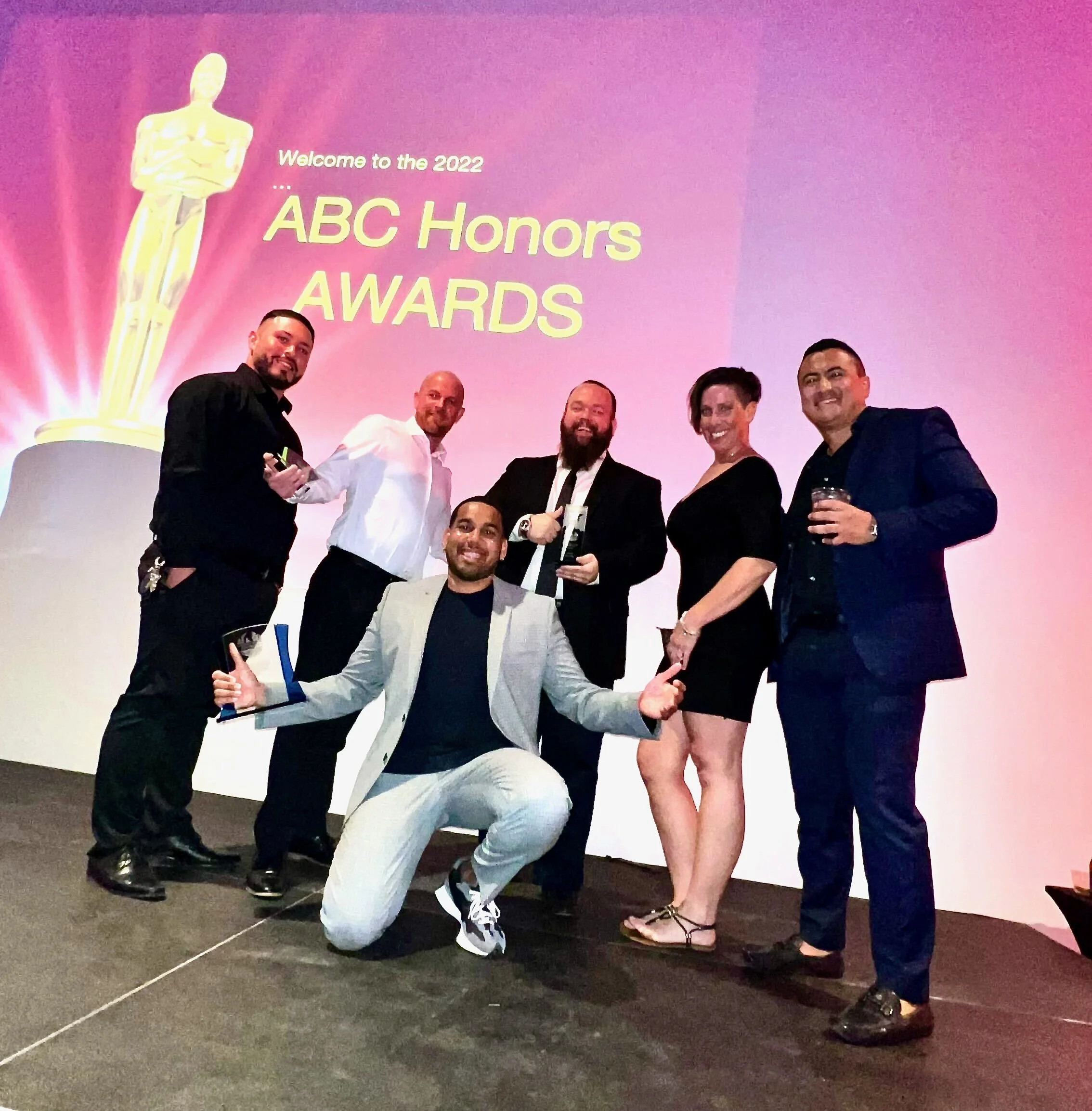 Group of six people celebrating at the ABC Honors Awards event, with a pink background and a large golden Oscar statue graphic.