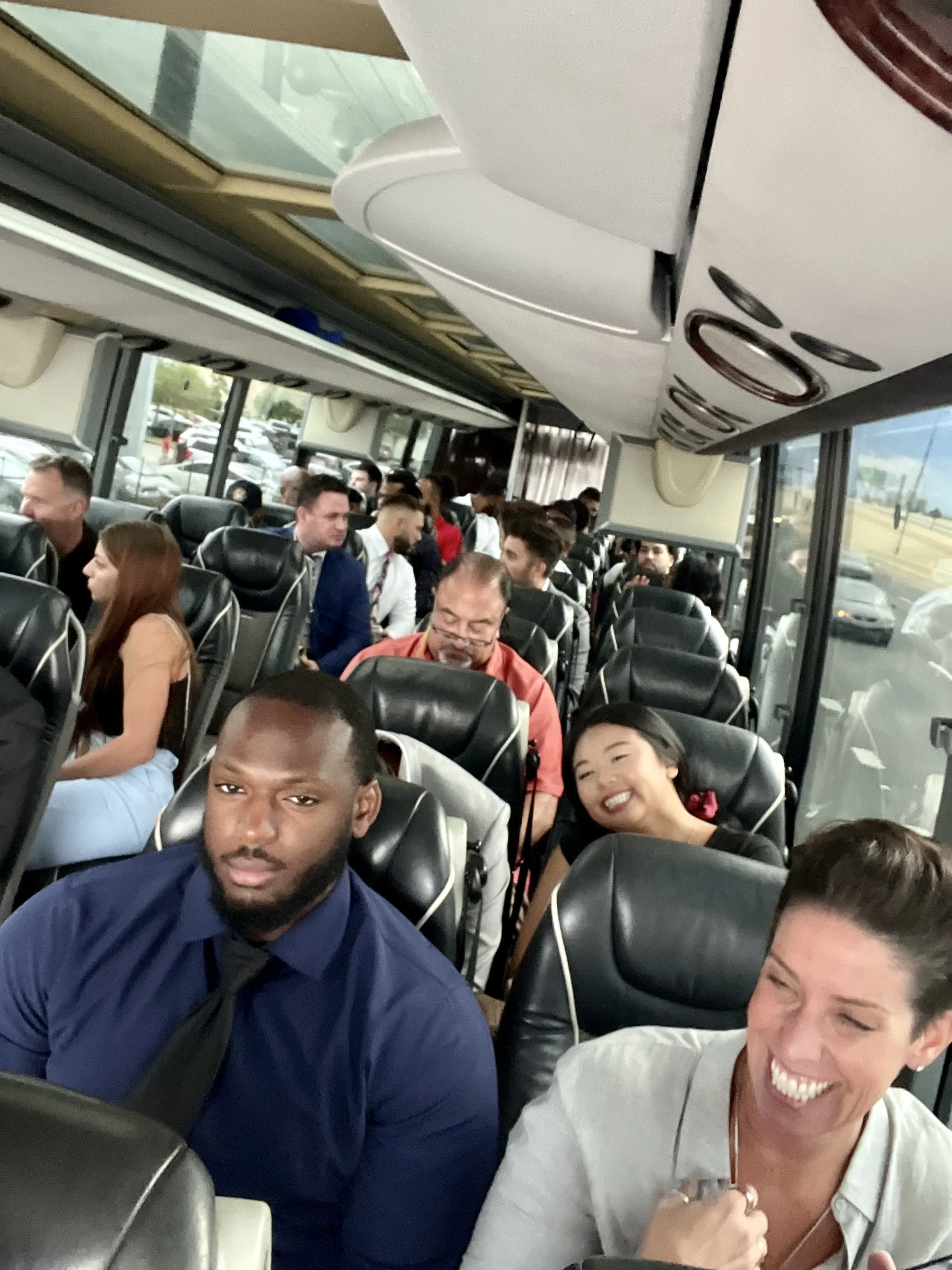 People sitting on a bus, some are smiling and chatting, and others are looking forward. The bus has large windows, with parked cars visible outside.