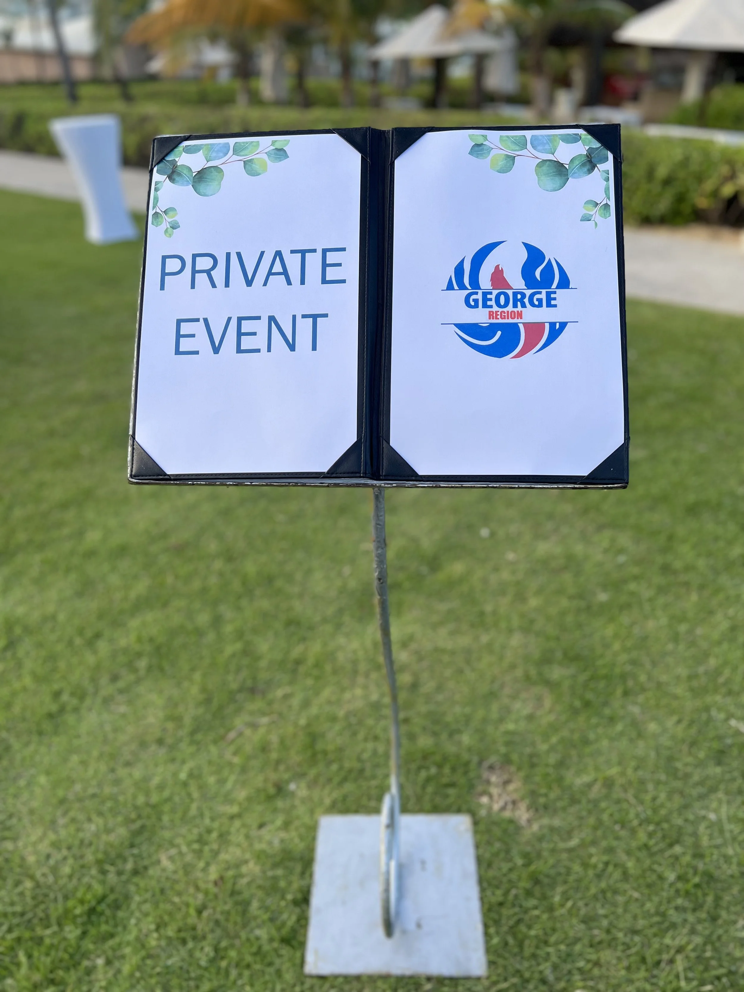 Sign with 'Private Event' on the left and a logo with 'George Region' on the right, placed outdoors on a grassy area with trees and structures in the background.