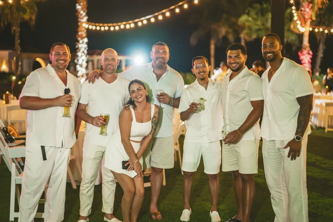 Group of friends enjoying an outdoor night party, dressed in white, holding drinks, with string lights and palm trees in the background.