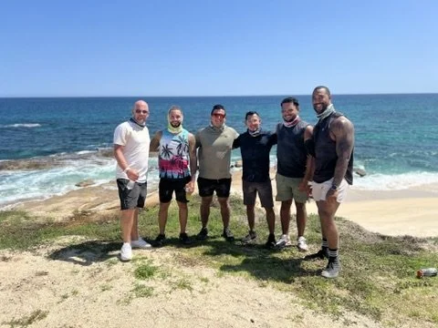 Six men standing together on a sandy beach near the water with waves, under a clear blue sky.