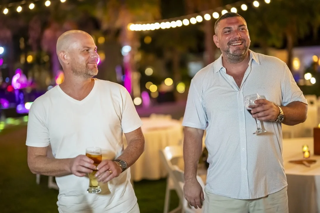 Two men standing outdoors at a nighttime event, each holding a drink and smiling, with string lights and decorated tables in the background.