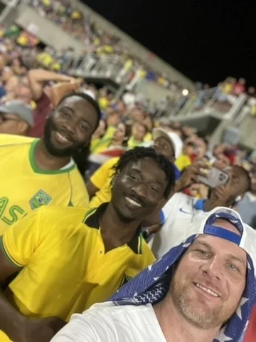 People smiling at a sports game, wearing yellow jerseys, in a stadium filled with spectators.