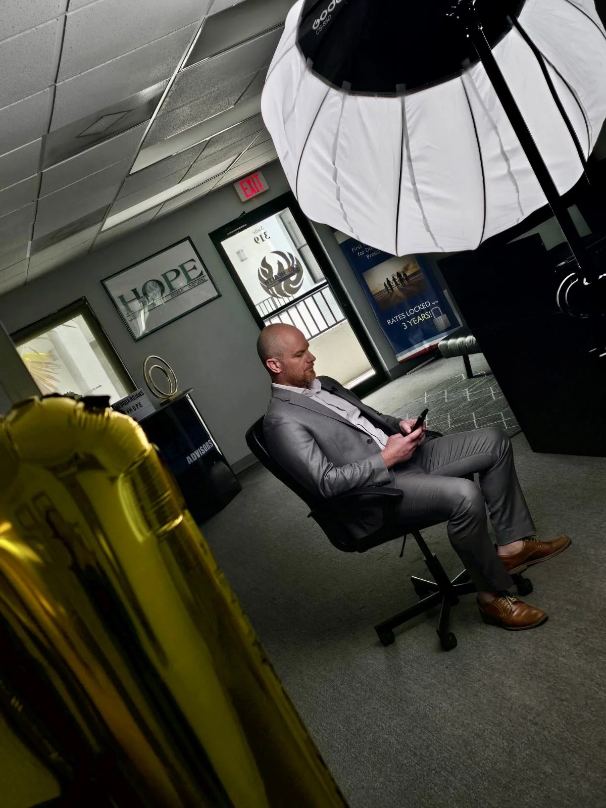 A man in a gray suit sitting on a black office chair, looking at his phone, in a modern office or waiting area with banners and signs on the wall.