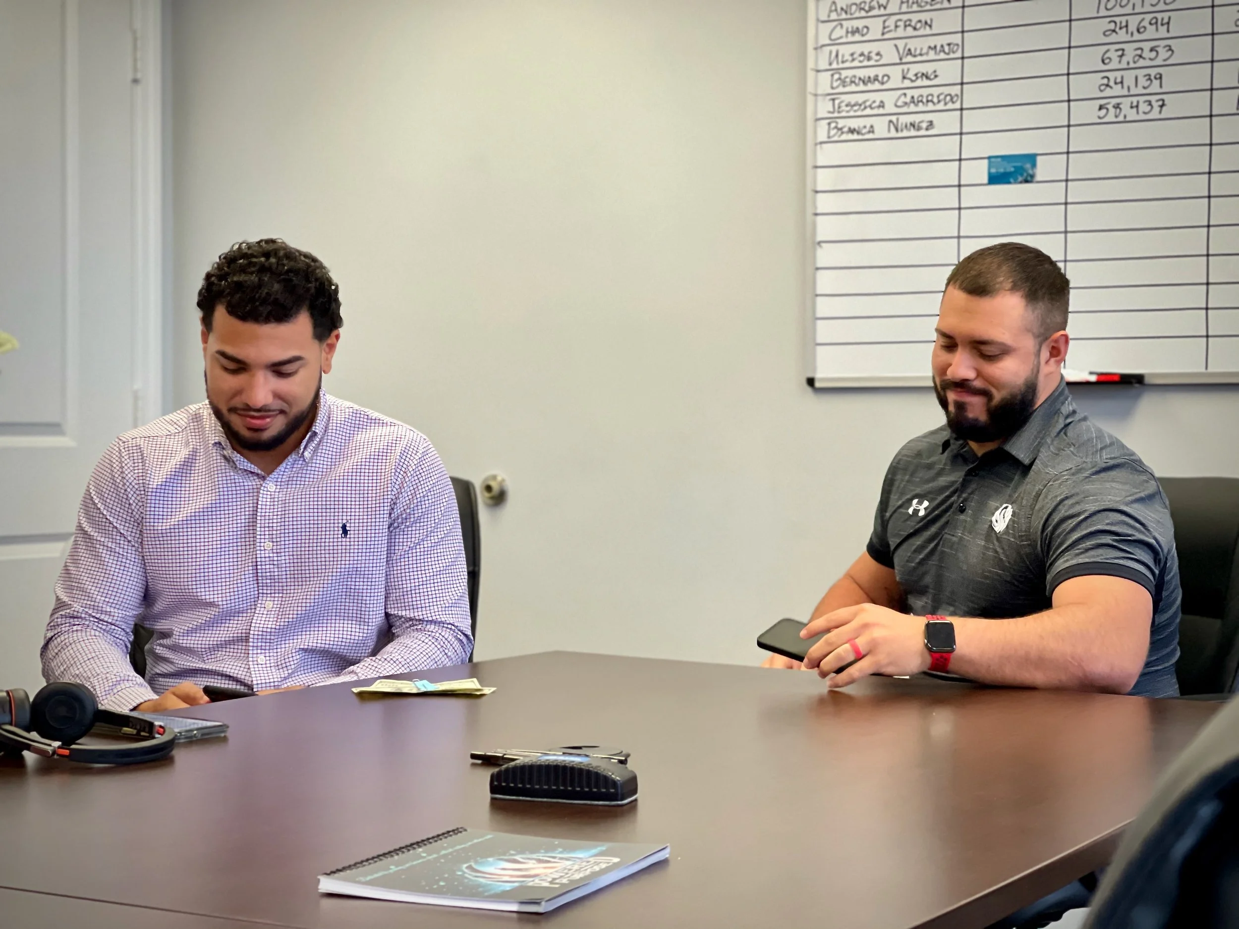 Two men sitting at a conference table, both looking at their phones. On the wall behind them is a whiteboard with a list of names and numbers.