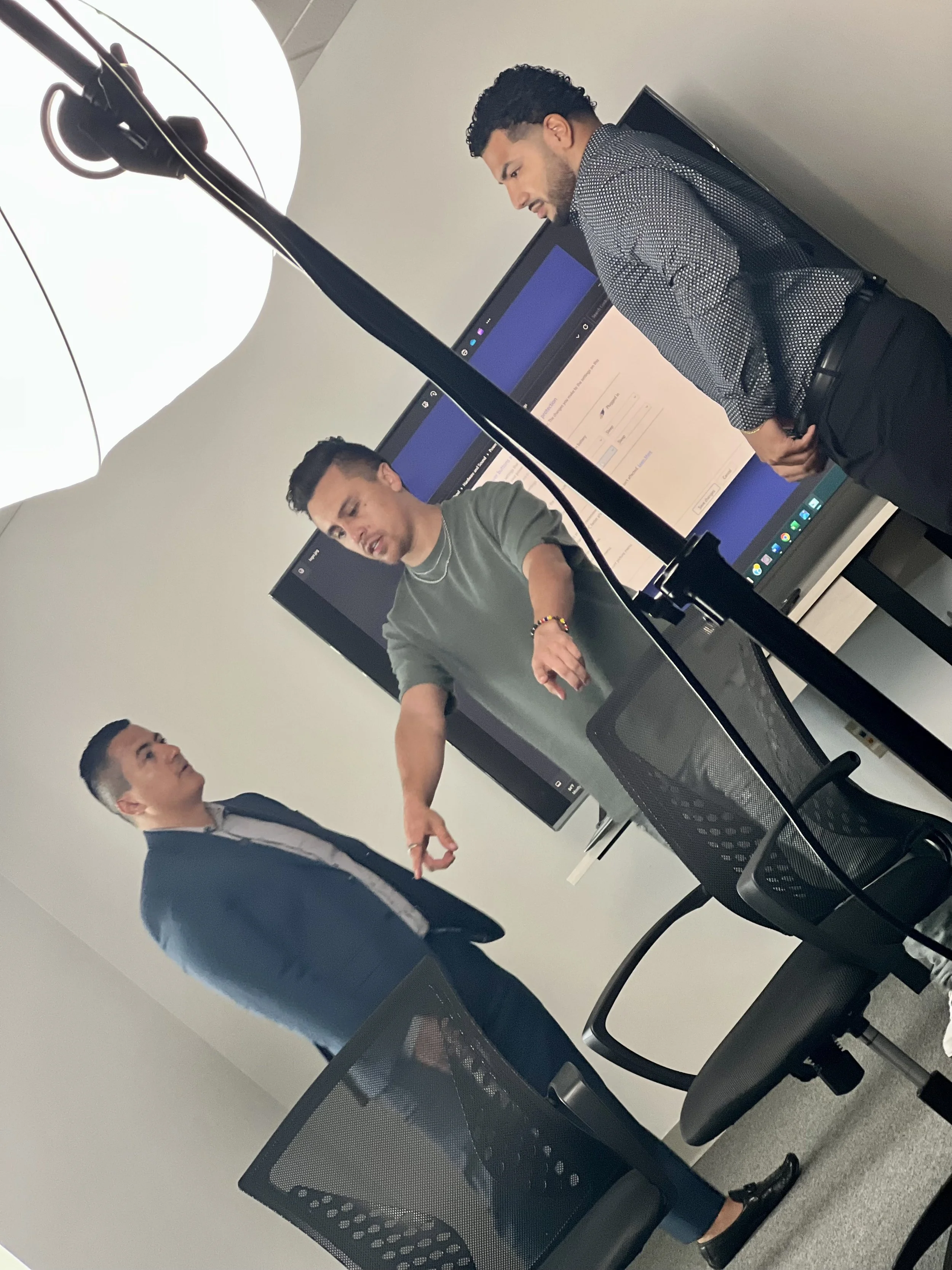 Three men in a business meeting or presentation in a conference room with two large screens and a conference table and chairs.