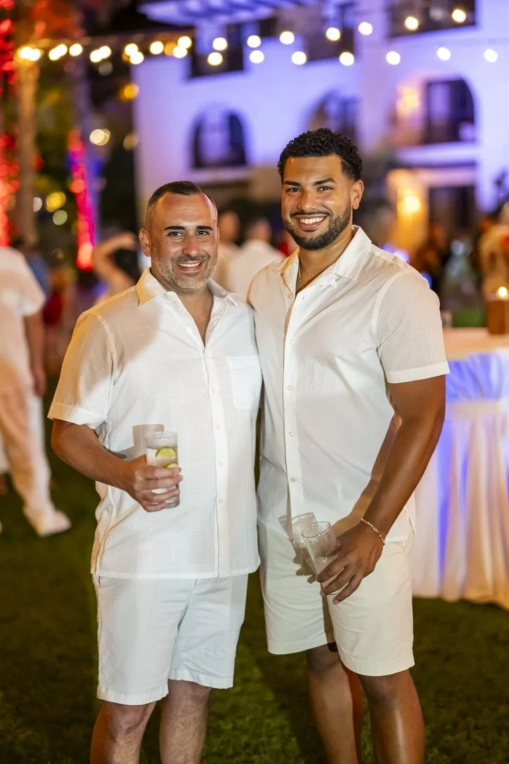 Two men smiling and standing close together at a nighttime outdoor event with string lights and a building in the background. Both are wearing white shirts and holding drinks.
