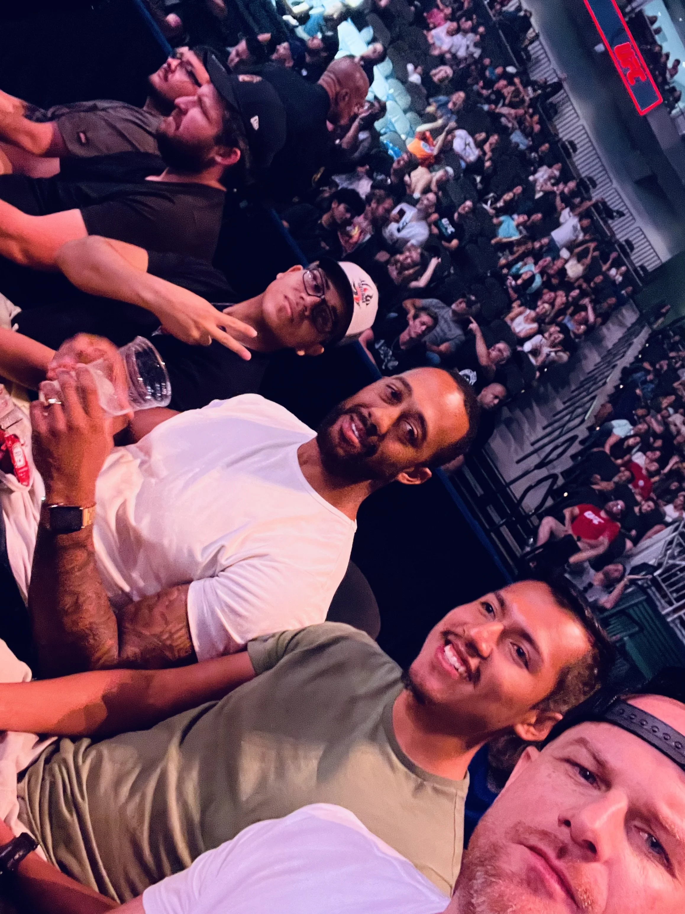 Four men sitting in a stadium, smiling and posing for a selfie, with a crowd of people in the background.