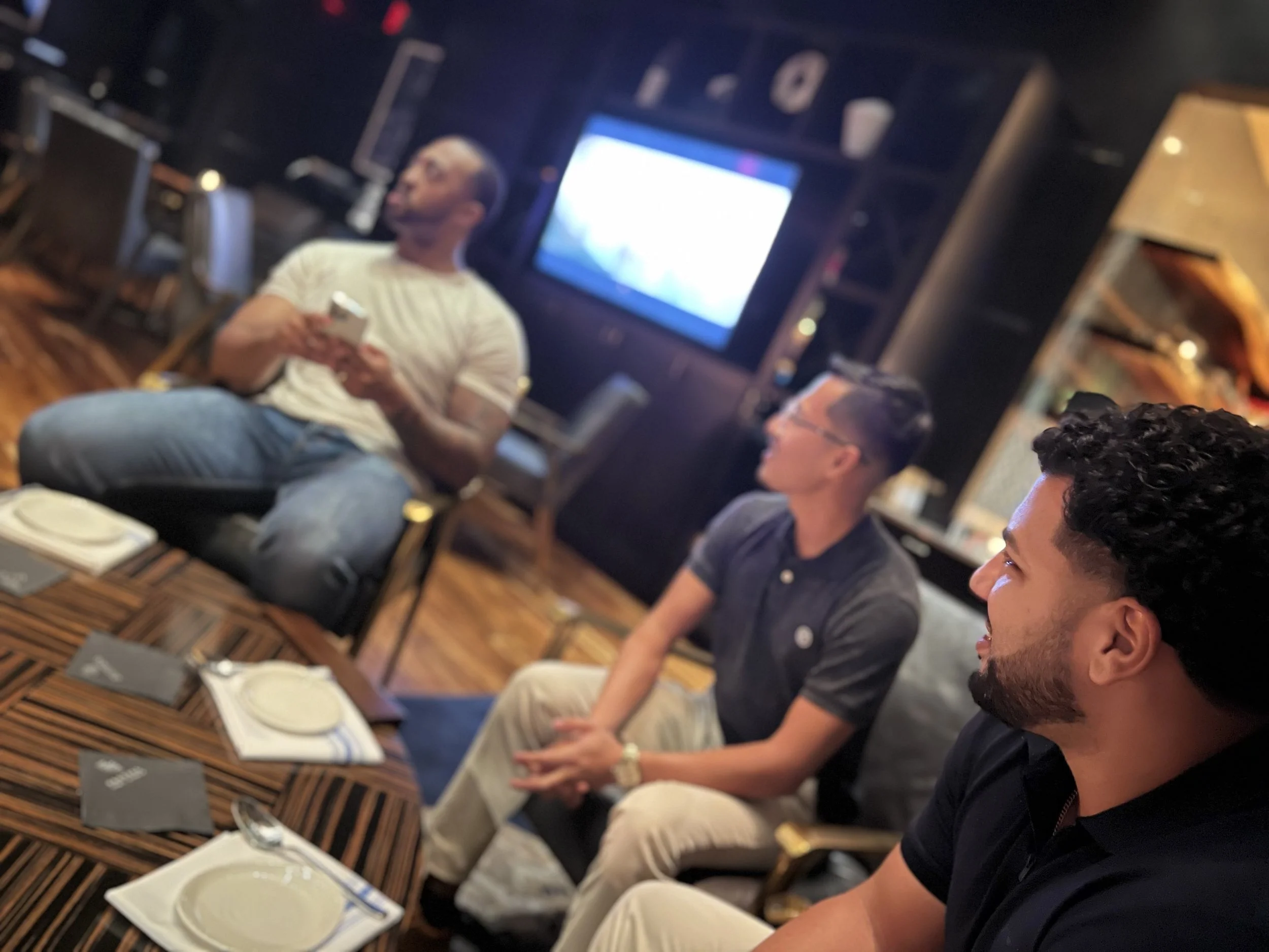 Three men seated at a table in a dimly lit restaurant or club, with a TV screen on the wall behind them, engaging in conversation or listening to someone out of frame.