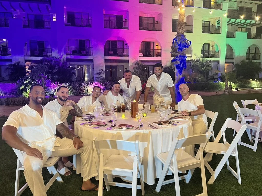 A group of seven men dressed in white, sitting and standing around a round table at an outdoor nighttime event with colorful building lights and palm trees in the background.
