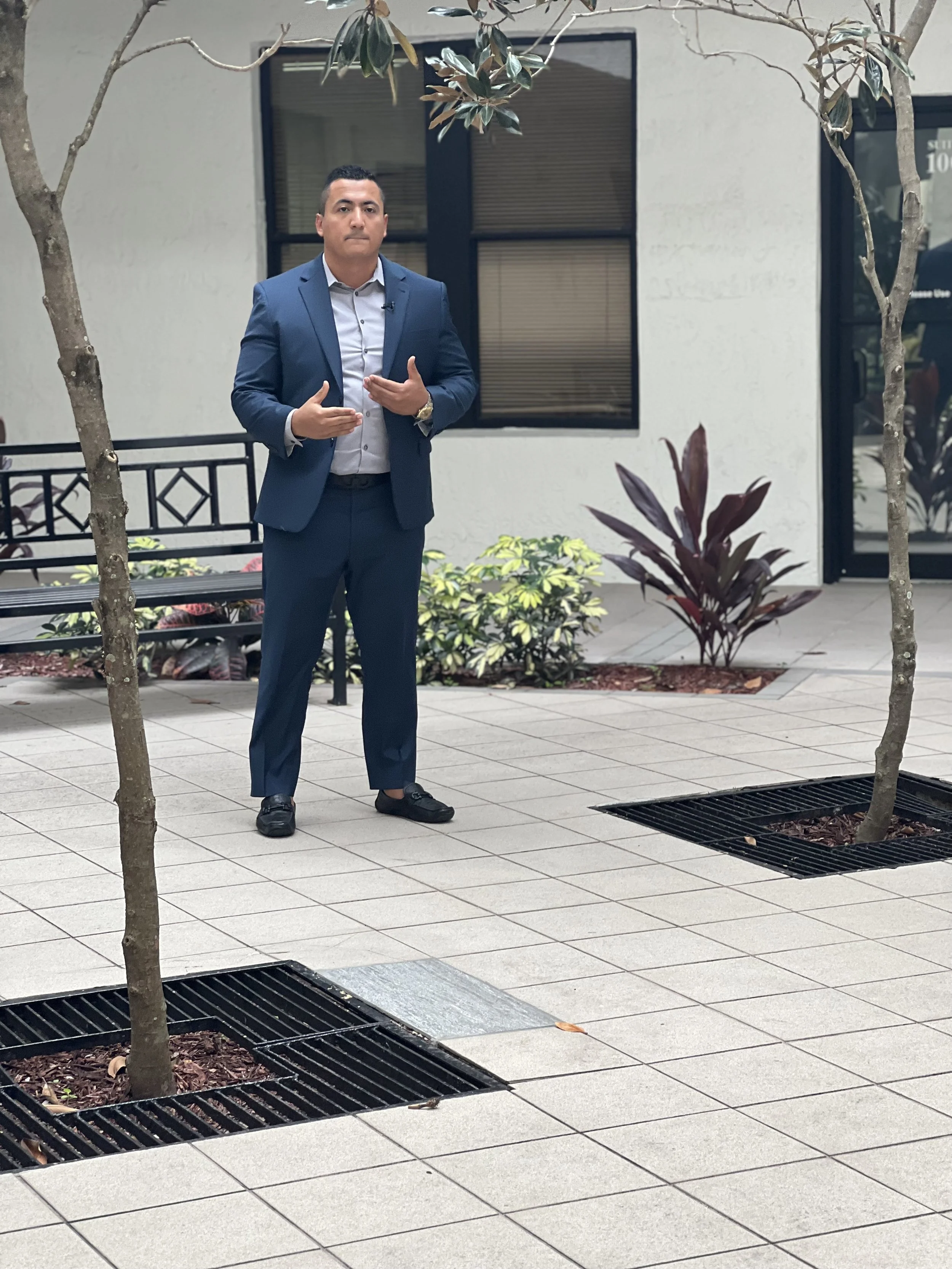 A man in a blue suit and white shirt standing outdoors on a tiled walkway, with trees and plants nearby, in front of a white building with windows.