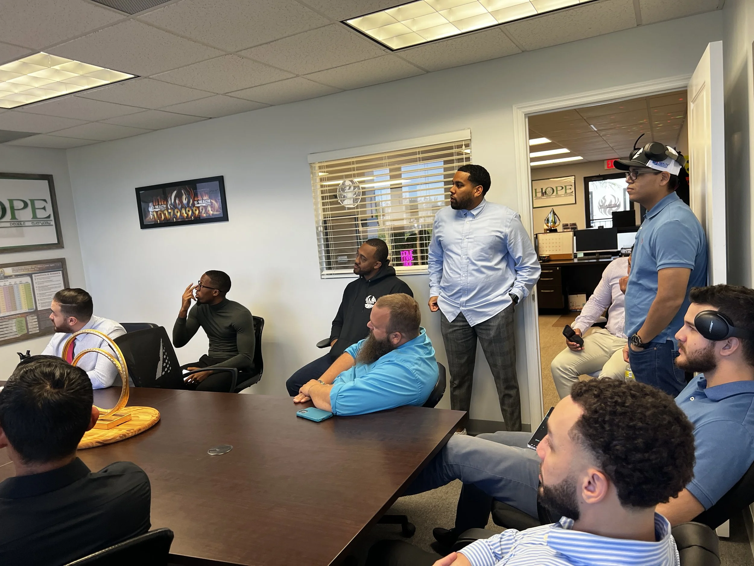Group of professionals attending a presentation in a conference room, some seated and some standing near the wall, with a man in a light blue shirt and checkered pants speaking or observing.