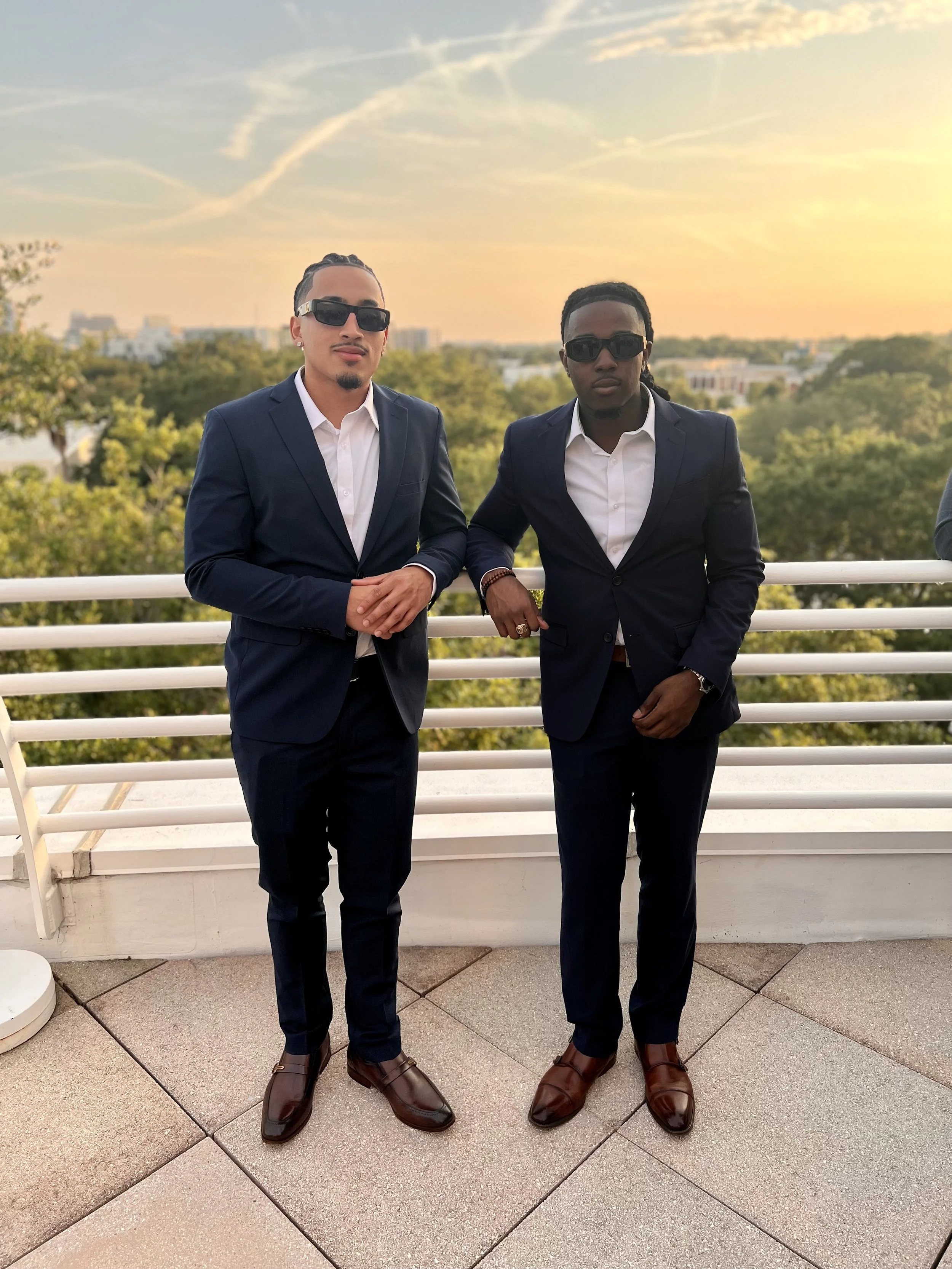Two men in dark blue suits and brown dress shoes standing on a balcony with a sunset view and green trees in the background.