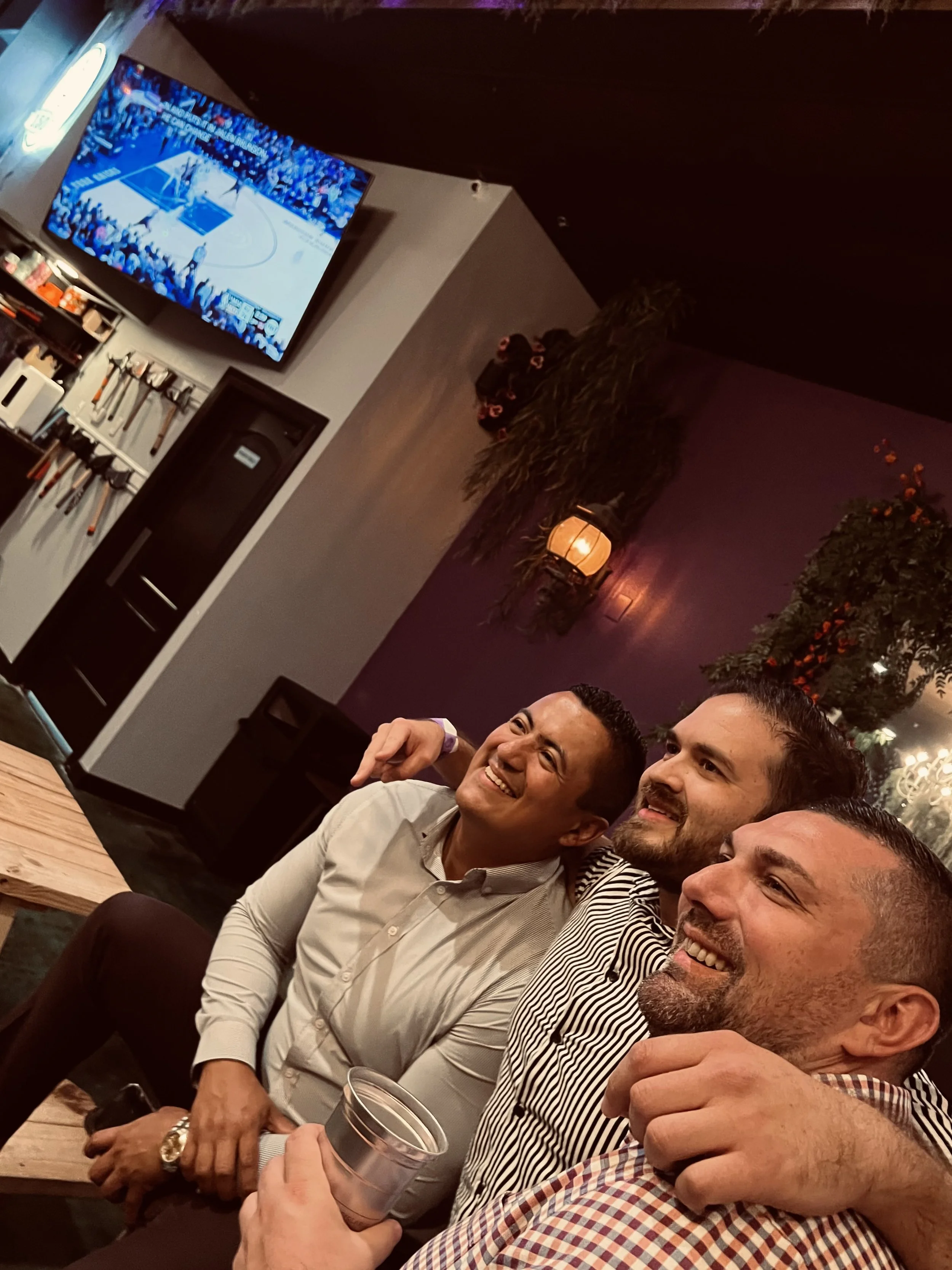 Three men smiling and sitting closely together, enjoying each other's company in a dimly lit bar or restaurant with a TV screen showing a basketball game in the background.