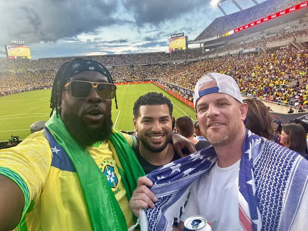 Three male fans at a soccer stadium, three cheers for Brazil, with the soccer field and crowd in the background.