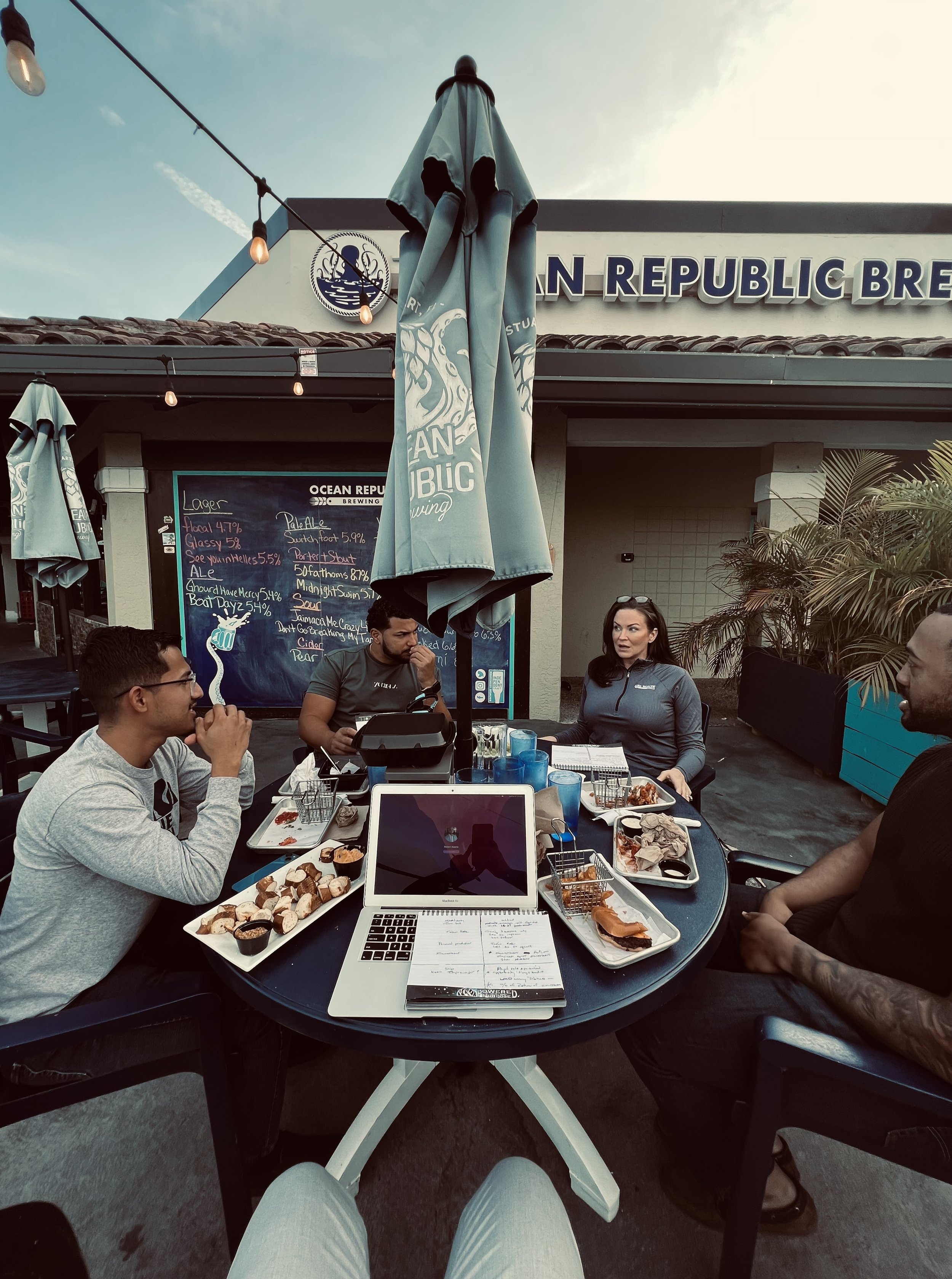 Four people sitting at an outdoor table at a brewery, with plates of food, drinks, and a laptop on the table, in front of a building with a sign that reads 'Ocean Republic Brewing' and a blackboard menu.