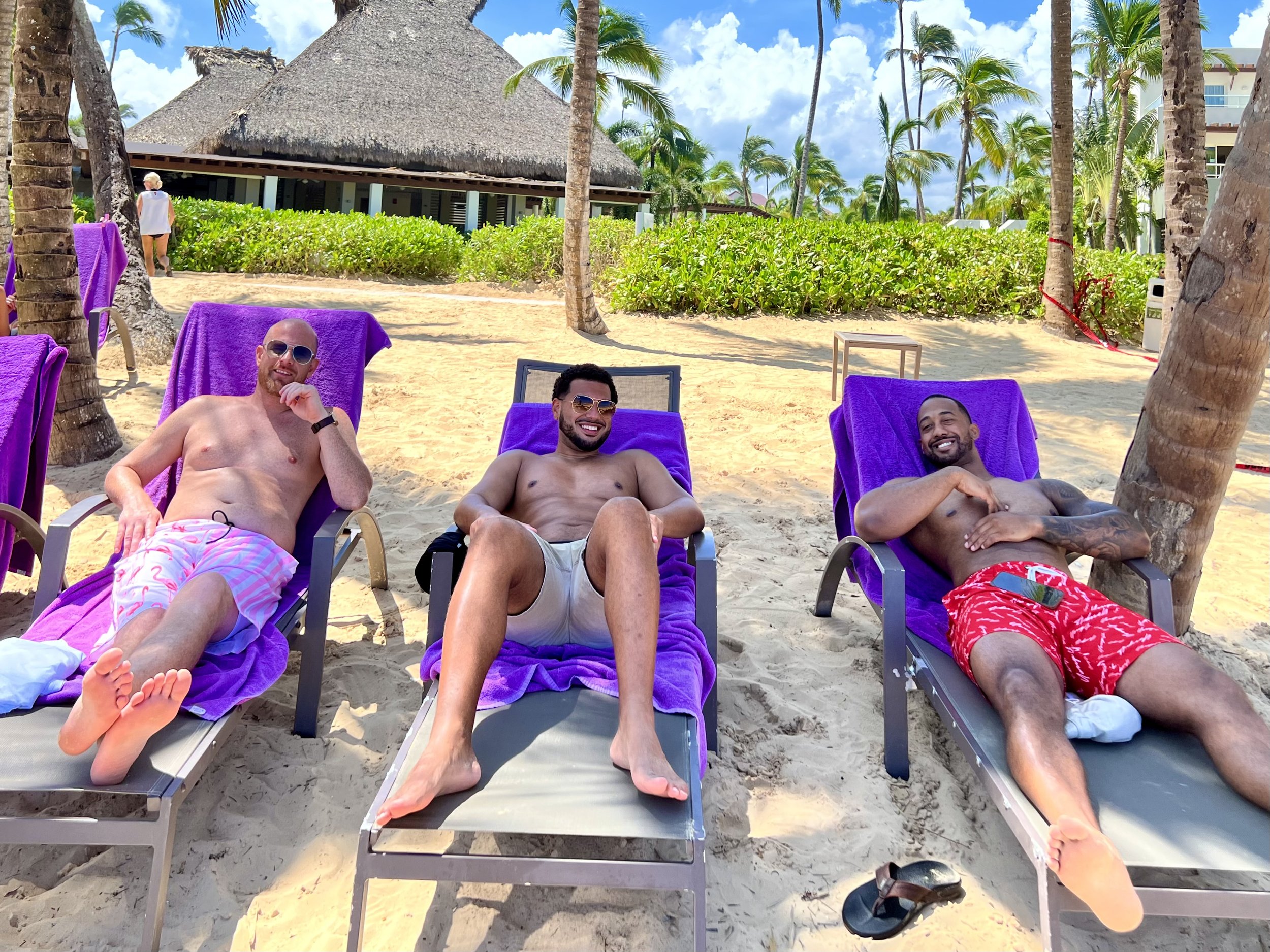 Three men relaxing on lounge chairs on a beach with purple towels, surrounded by palm trees, sand, and a thatched-roof hut in the background.