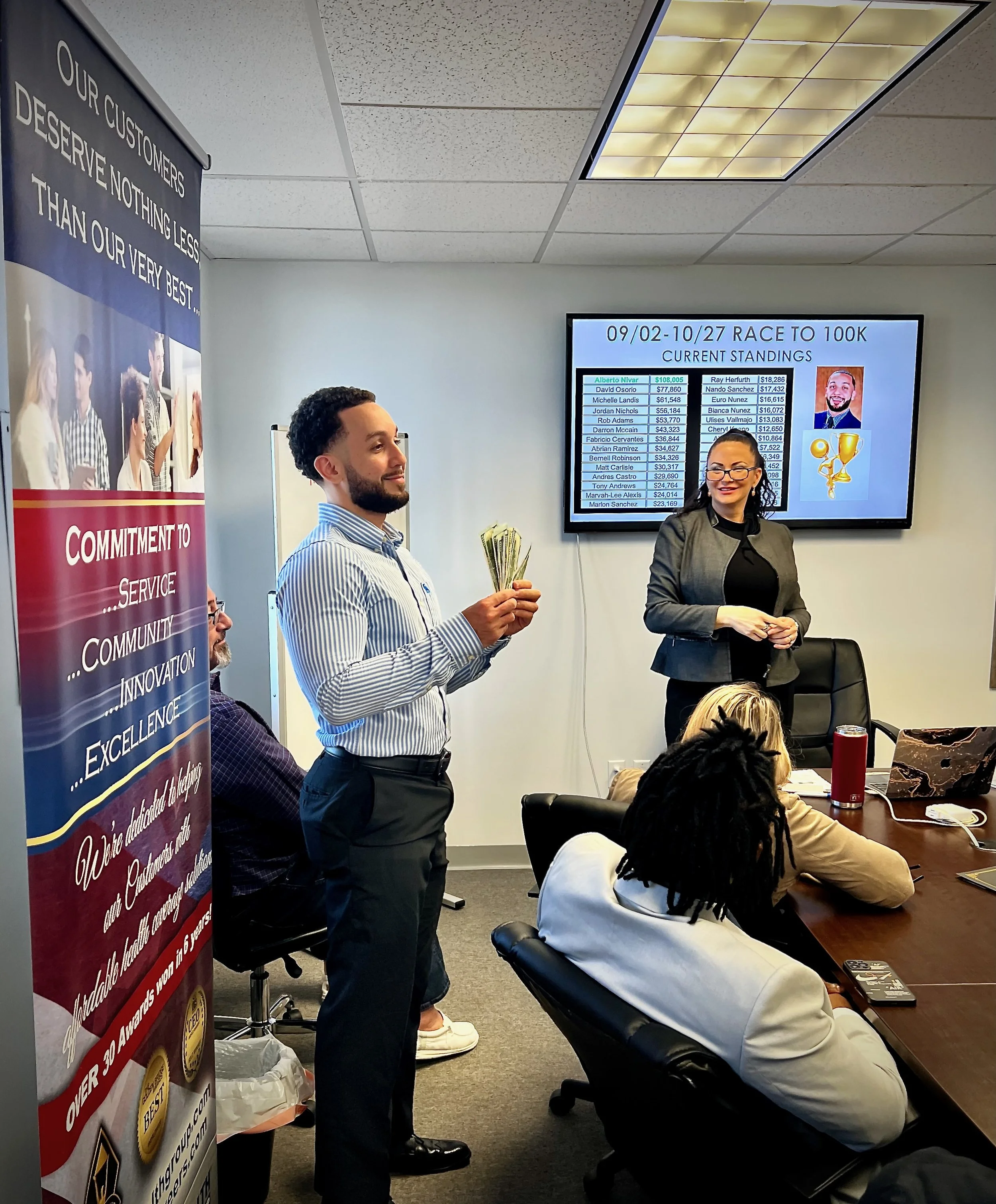 Man holding cash in a conference room during a meeting, with a woman standing at the front near a screen displaying race standings, and seated individuals at the table.