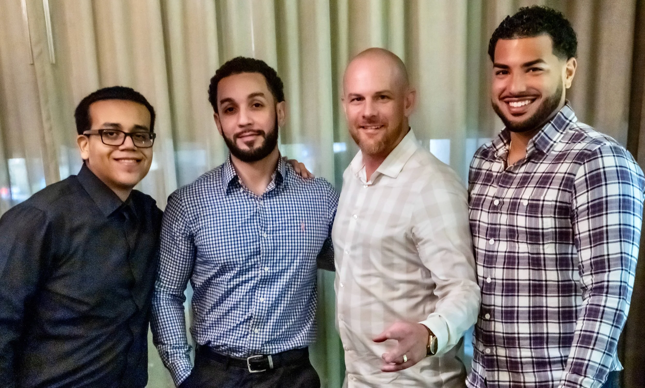 Four men standing together indoors in front of curtains, smiling for the photo.