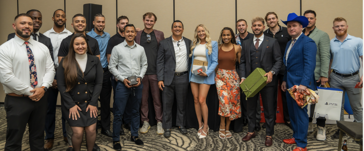 Group of professionally dressed young adults posing together at an indoor event, some holding wrapped gifts and boxes.