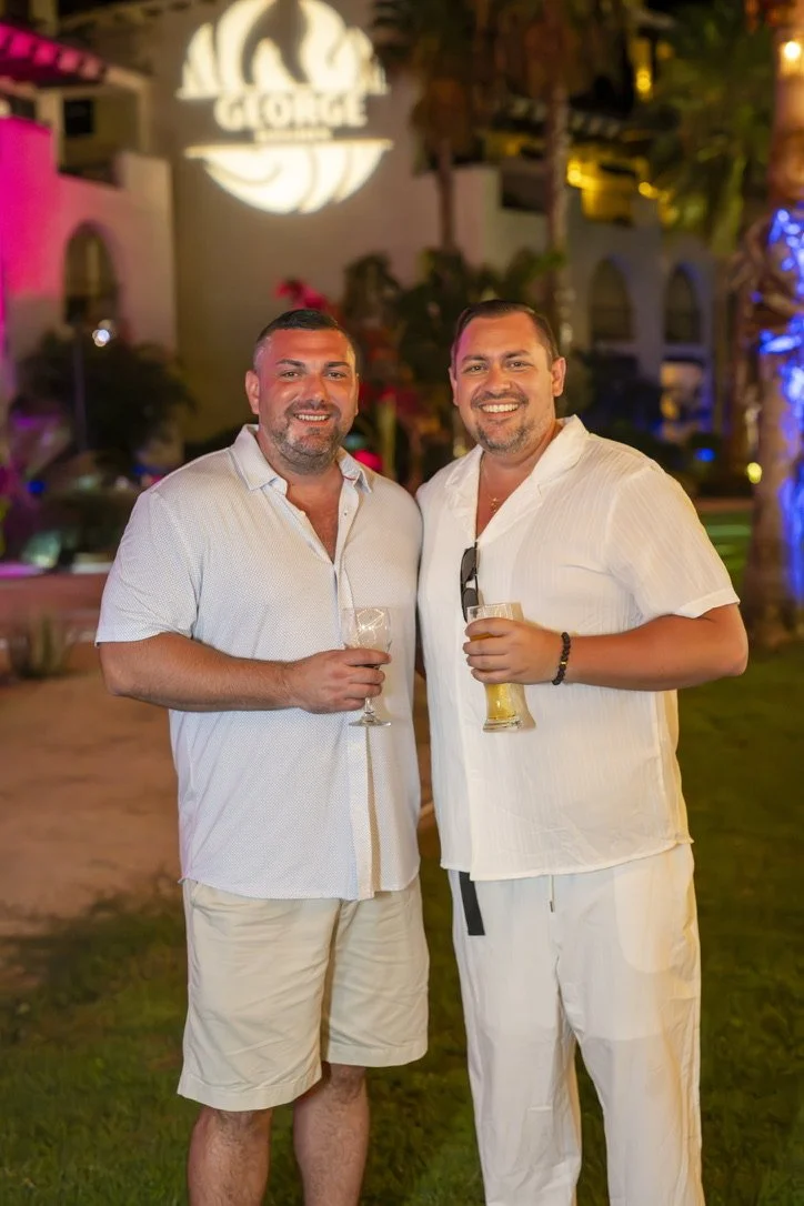 Two men smiling and holding drinks at an outdoor nighttime event. The background features colorful lighting, palm trees, and a large sign with the logo 'Gorge' illuminated above.