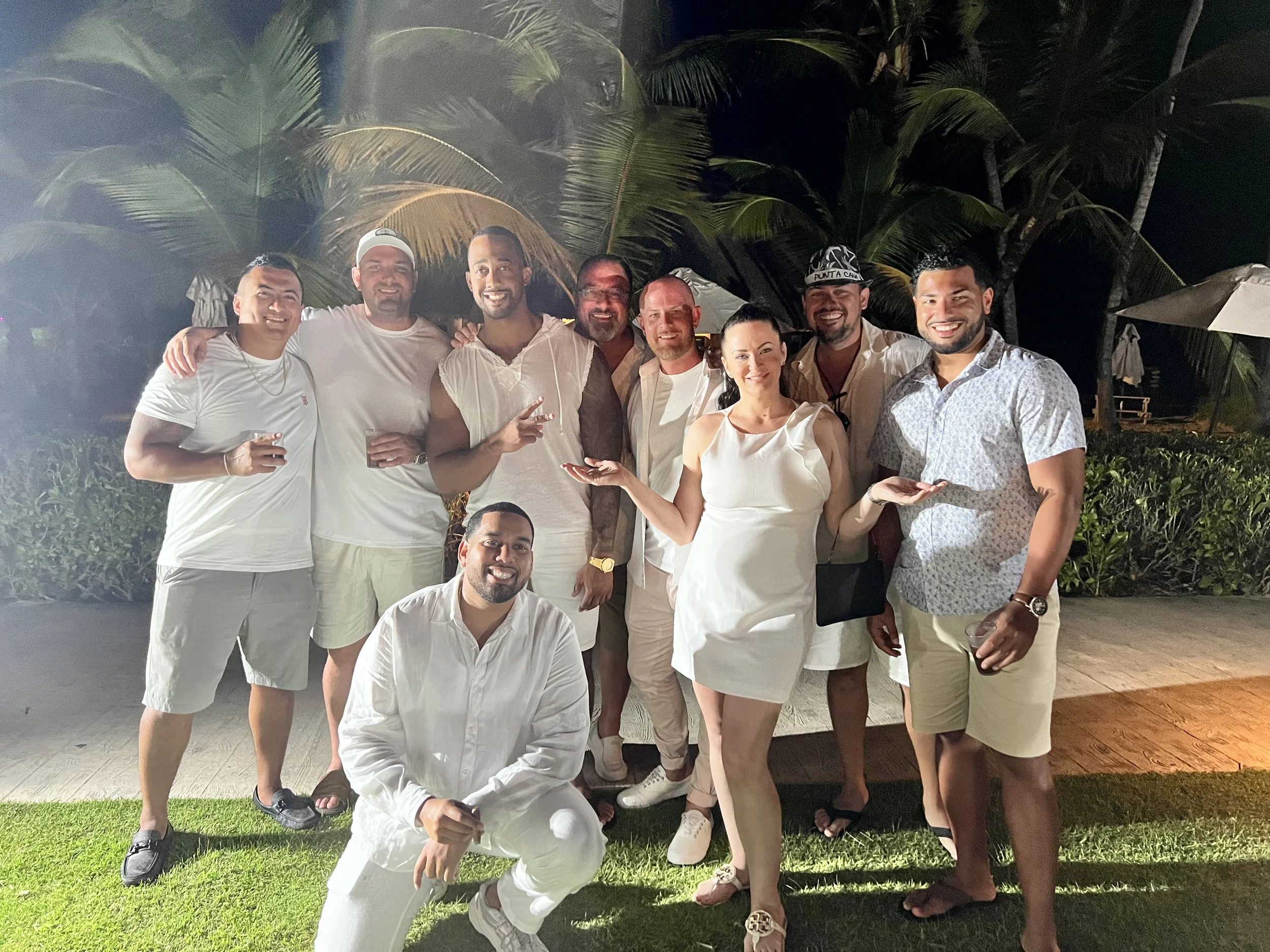 Group of eleven friends, dressed in casual white and light-colored clothes, smiling and posing at night outdoors on a patio with palm trees in the background.